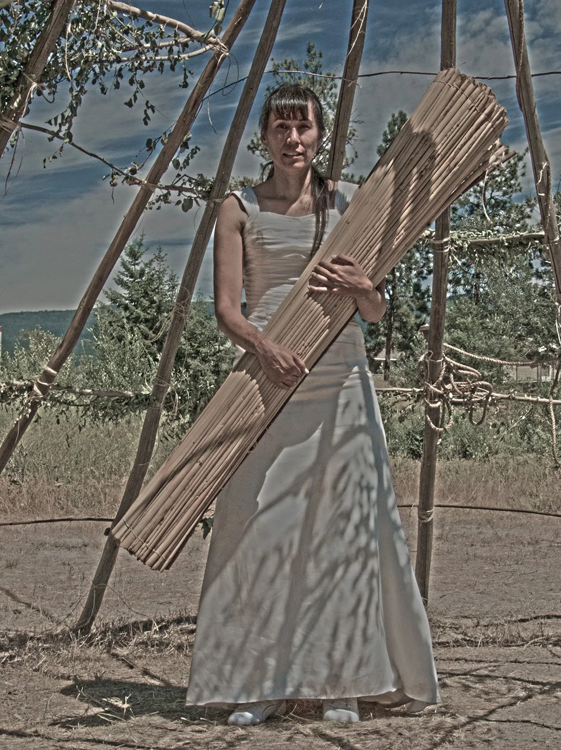 A woman standing outdoors, holding a large bundle of wooden sticks, with a makeshift triangular wooden structure behind her and trees in the background.