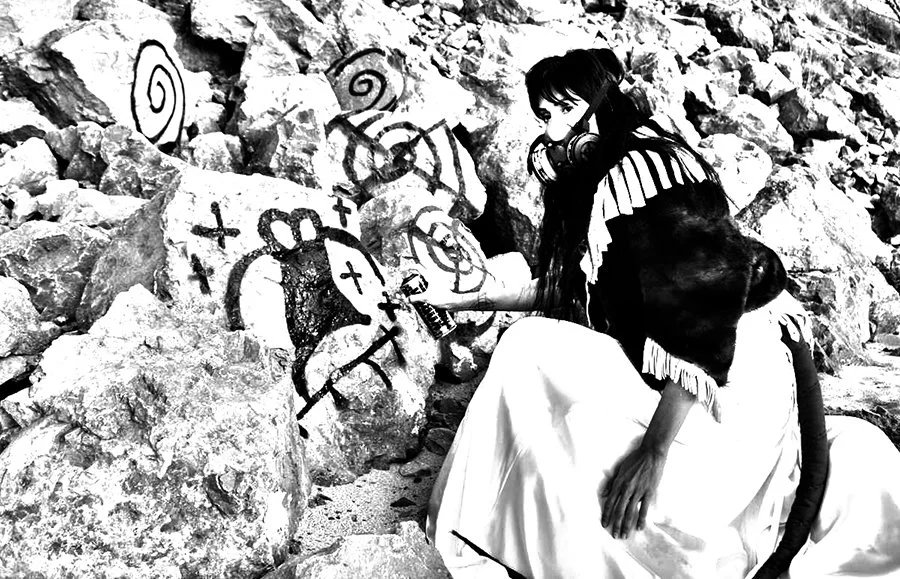 Woman dressed in a long skirt and shawl sitting on rocks in front of graffiti on stones in a rocky outdoor setting.