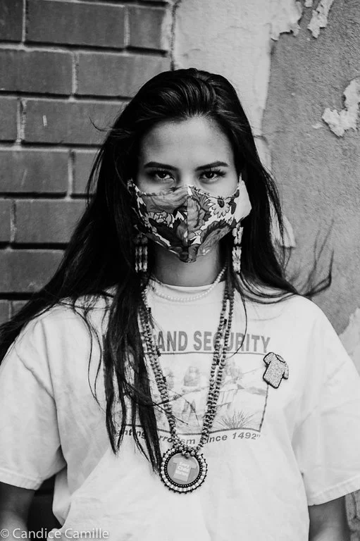 Black and white photo of a young woman wearing a floral face mask, earrings, a graphic T-shirt, and layered necklaces, standing against a textured wall with brick and peeling paint.