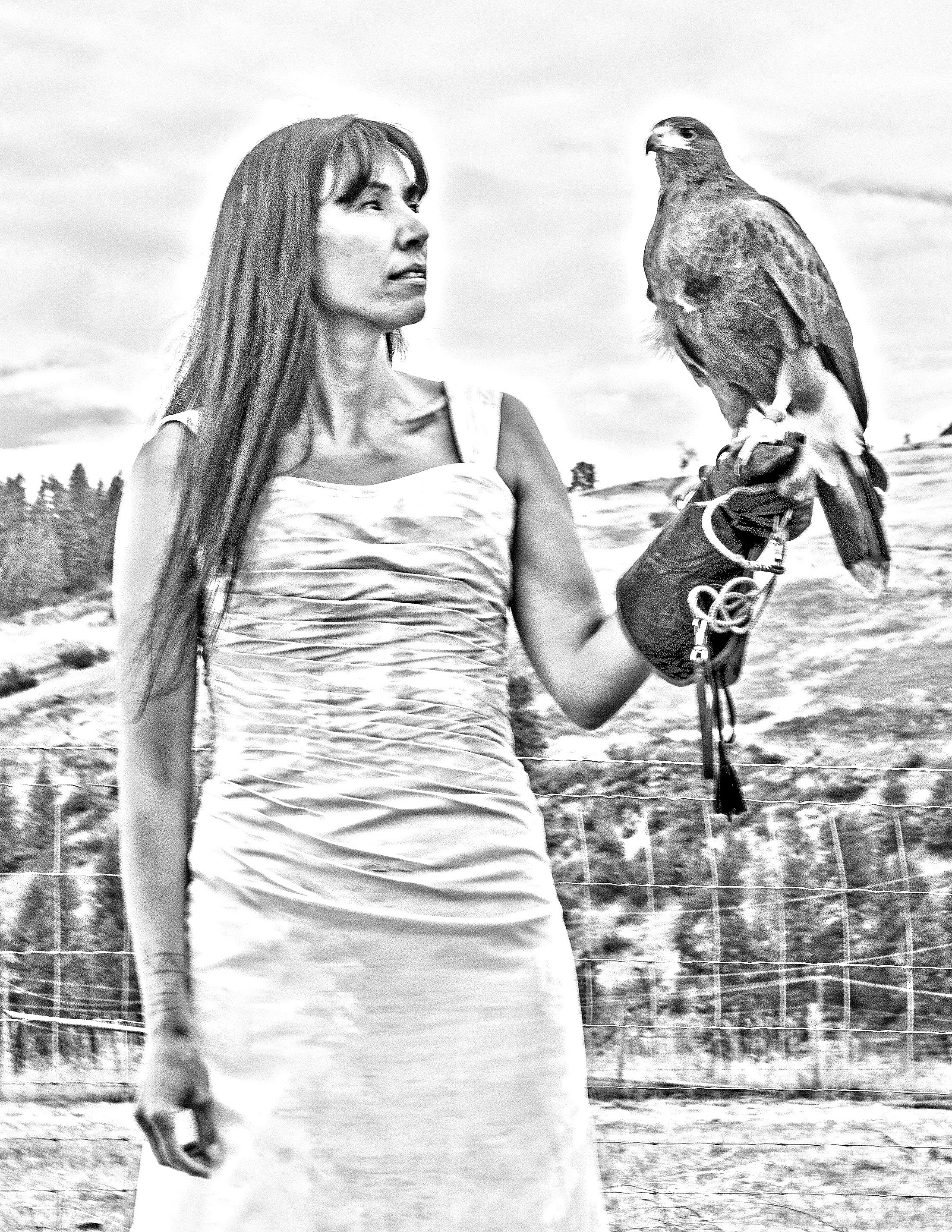 A woman holding a bird of prey on her gloved hand outdoors, with a fenced area and trees in the background.
