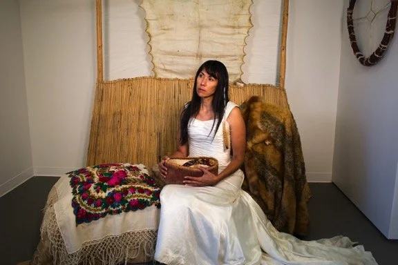 A woman in a white dress sitting on a vintage-style couch with a colorful crocheted blanket and a fur coat draped over her shoulder, holding a basket in her lap, in an art gallery or museum setting.