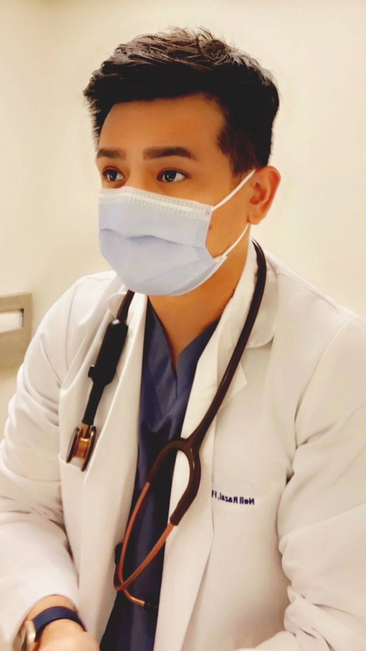 A young male doctor wearing a face mask, white lab coat, and stethoscope around his neck, sitting in a medical facility.