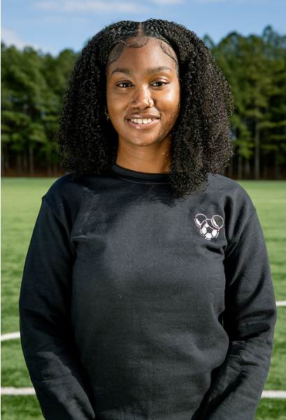 Young woman with curly black hair smiling outdoors on a grassy field with trees in the background, wearing a black sweatshirt with a small soccer ball graphic