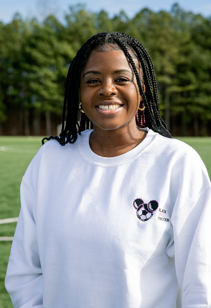 A smiling woman with braided hair standing outdoors on a grassy field with trees in the background, wearing a white sweatshirt with a black and white logo and the text "BLACK SOCCER."