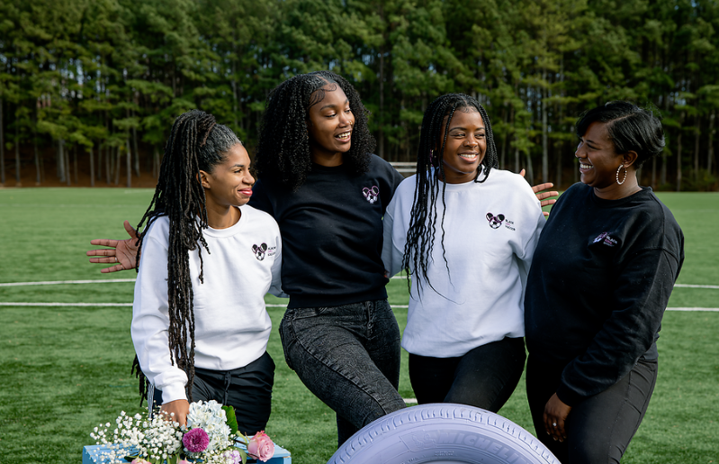 Four women standing on a sports field, smiling and laughing together, with trees in the background. Standing in front of a bouquet of flowers