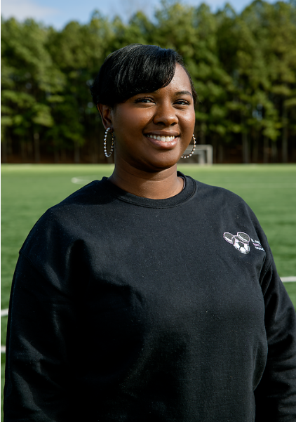 A woman standing outdoors on a grassy field with trees in the background, smiling at the camera, wearing earrings and a black sweatshirt.