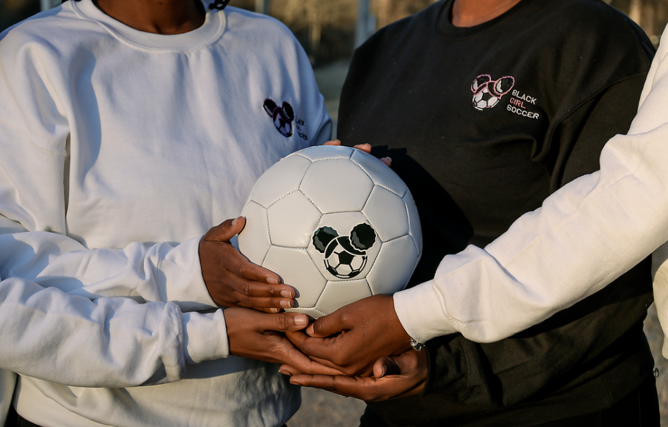 Three people holding a soccer ball with panda face graphics, wearing black and white 'Black Girl Soccer' shirts, outdoors.