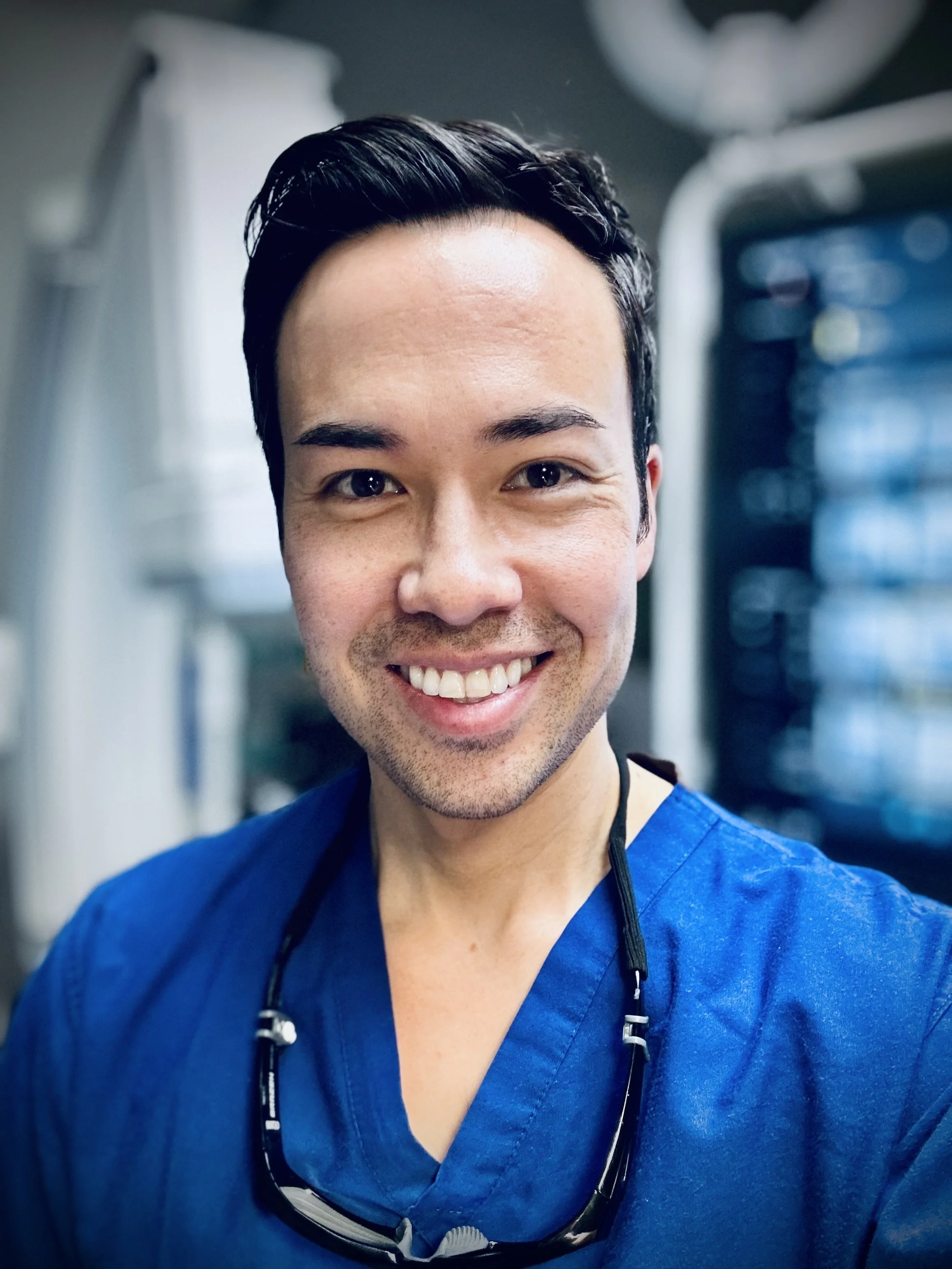 A smiling man with short dark hair wearing blue medical scrubs and black glasses hanging around his neck in a clinical setting.