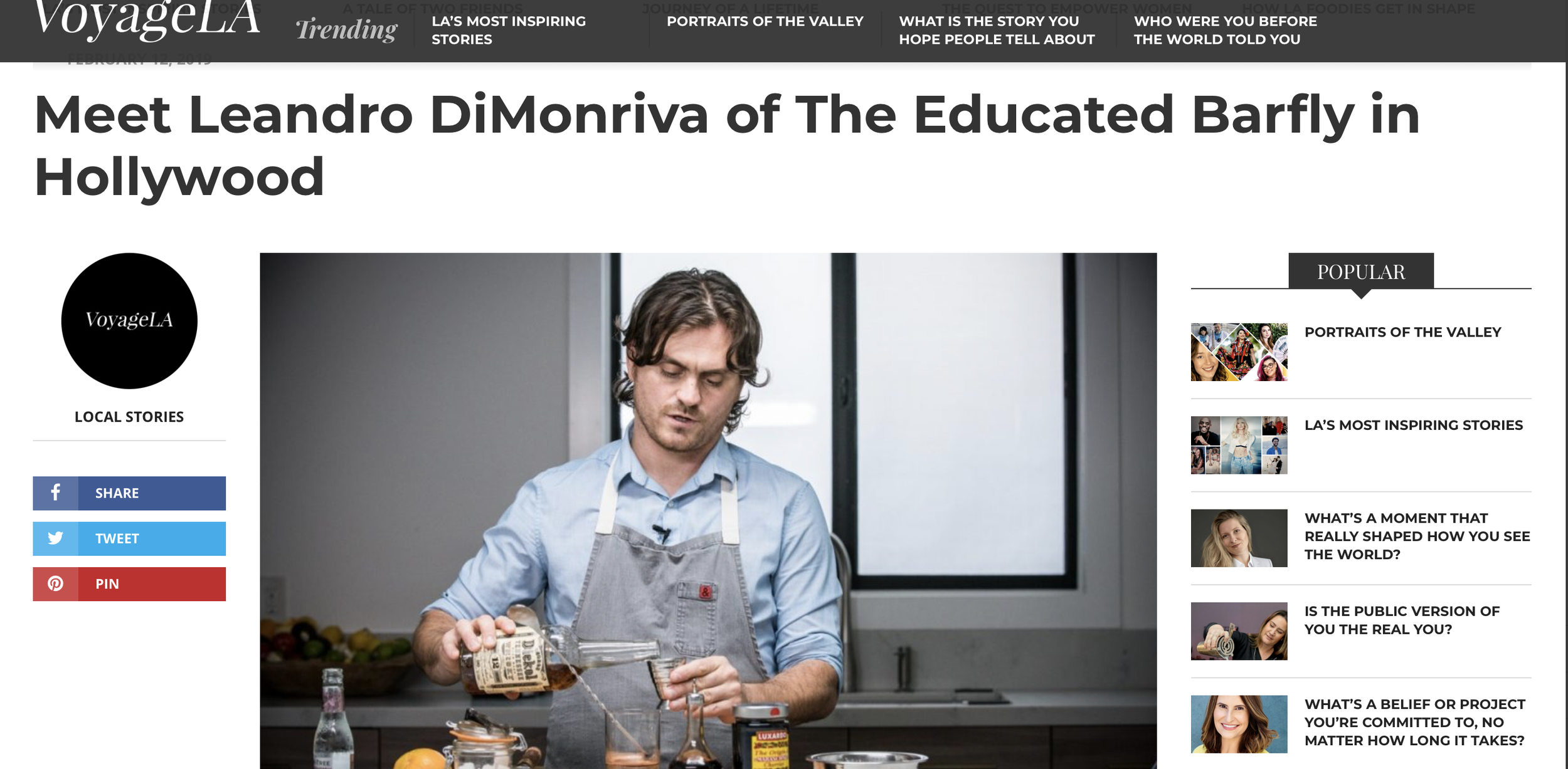 A man with curly brown hair, wearing a blue shirt and gray apron, pouring a beverage into a glass in a modern kitchen. There is a window behind him and various bottles and ingredients on the counter.