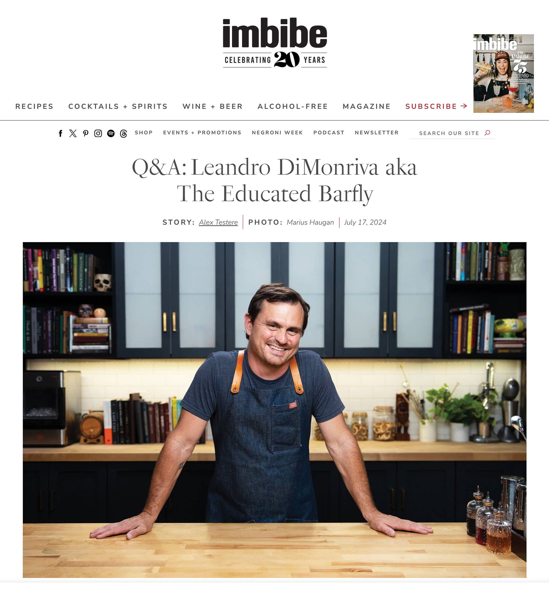 A man in a kitchen wearing a denim apron, standing and leaning on a wooden counter, smiling at the camera with dark brown hair, dark shirt, and in front of shelves filled with books, a skull, and kitchen items.