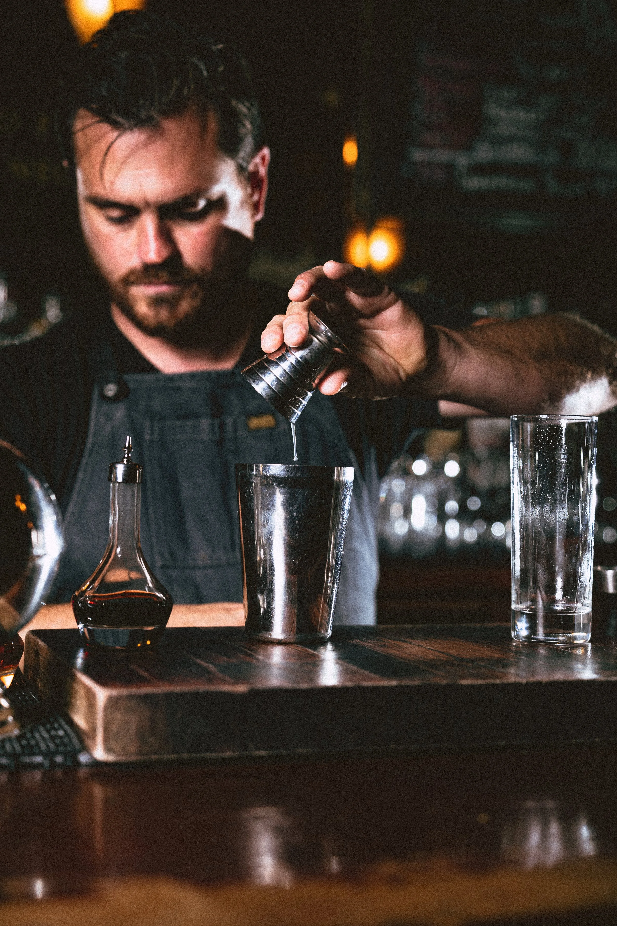 Bartender pouring a drink into a metal shaker in a dimly lit bar.