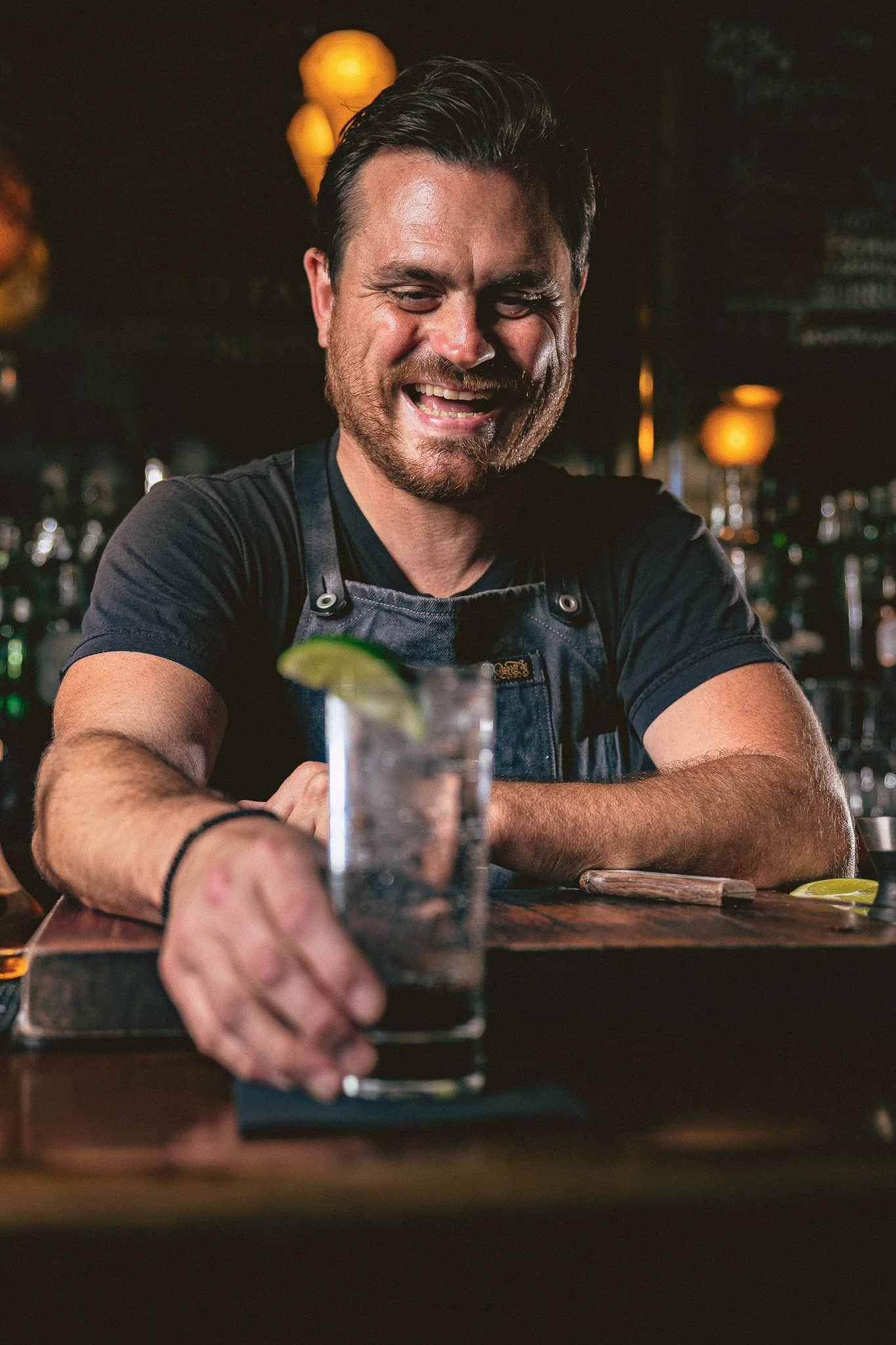 A smiling bartender with dark hair and a beard serving a drink at a bar, with a lime wedge on the glass.