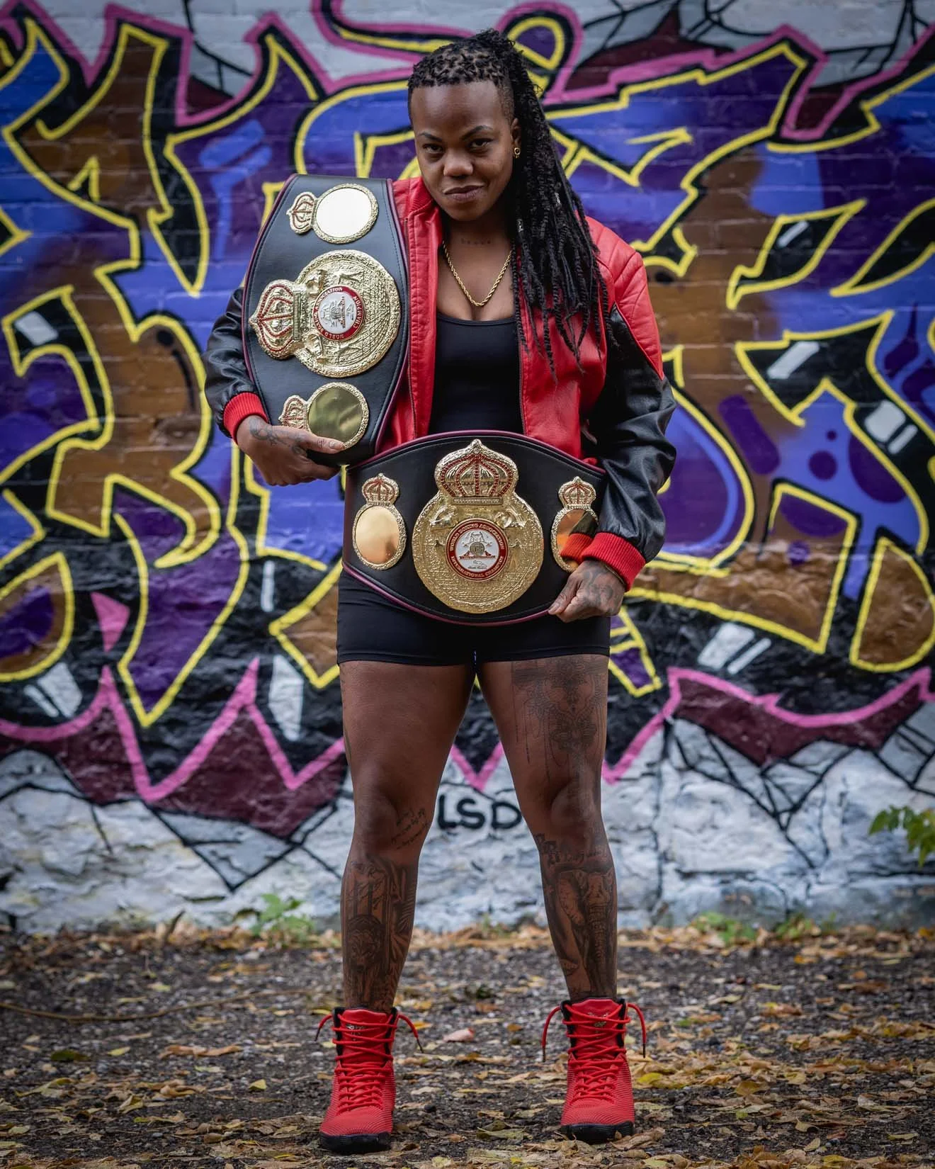 A woman with long dreadlocks holding two championship belts, standing in front of a colorful graffiti wall. She is wearing a black dress, red and black jacket, red boots, and has tattoos on her legs. She has a confident expression on her face.