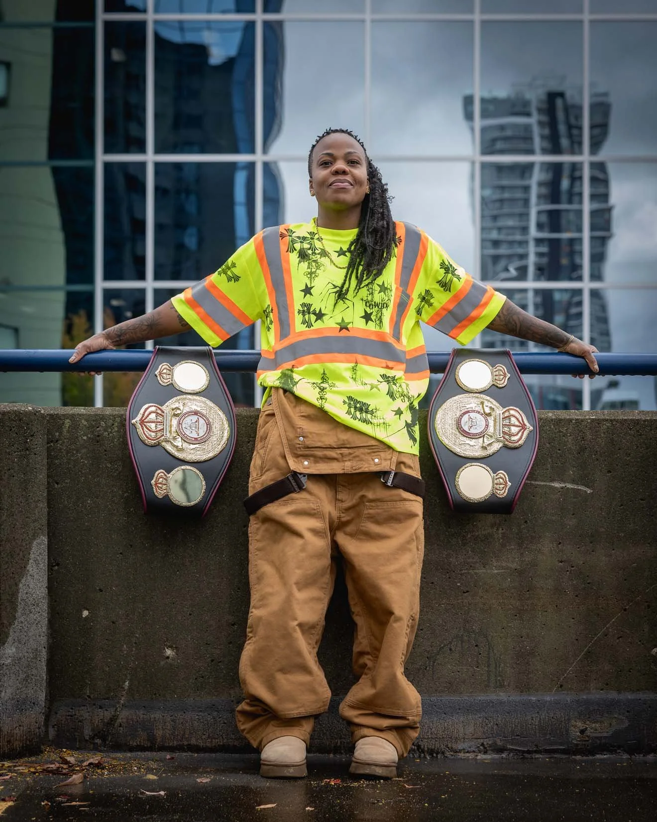 A person standing outdoors in front of a glass building, wearing a high-visibility shirt with reflective stripes, loose brown pants, beige shoes, and holding two championship belts with gold plates. The person's arms are outstretched and they are smi