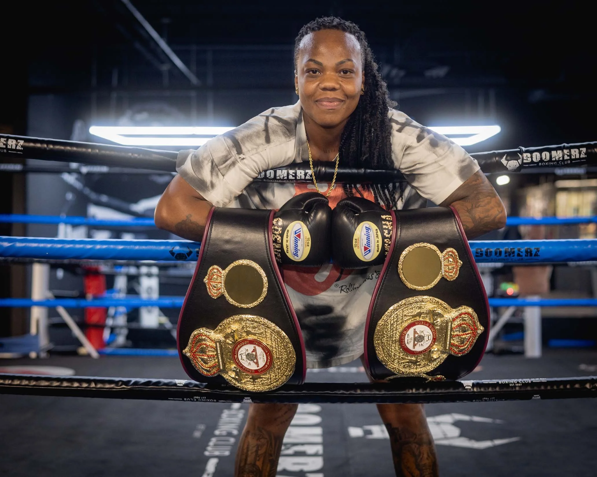 A female boxer is in the boxing ring, leaning on the ropes with her arms, wearing boxing gloves and multiple championship belts over her waist, on a dark background with bright lights overhead.