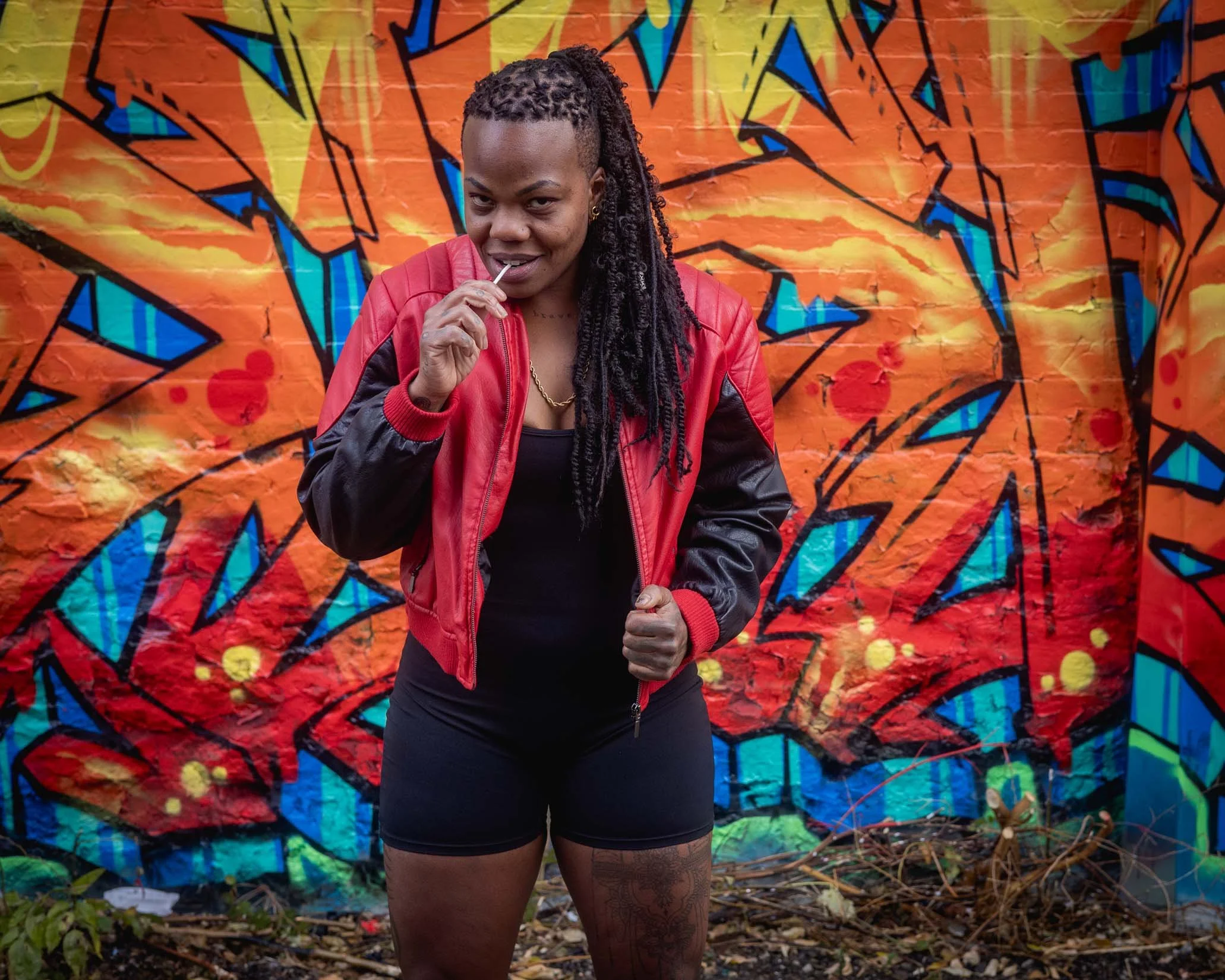 A woman with dreadlocks wearing a red jacket and black shorts, standing outdoors in front of a colorful graffiti wall, holding a lollipop in her mouth and making a fist.