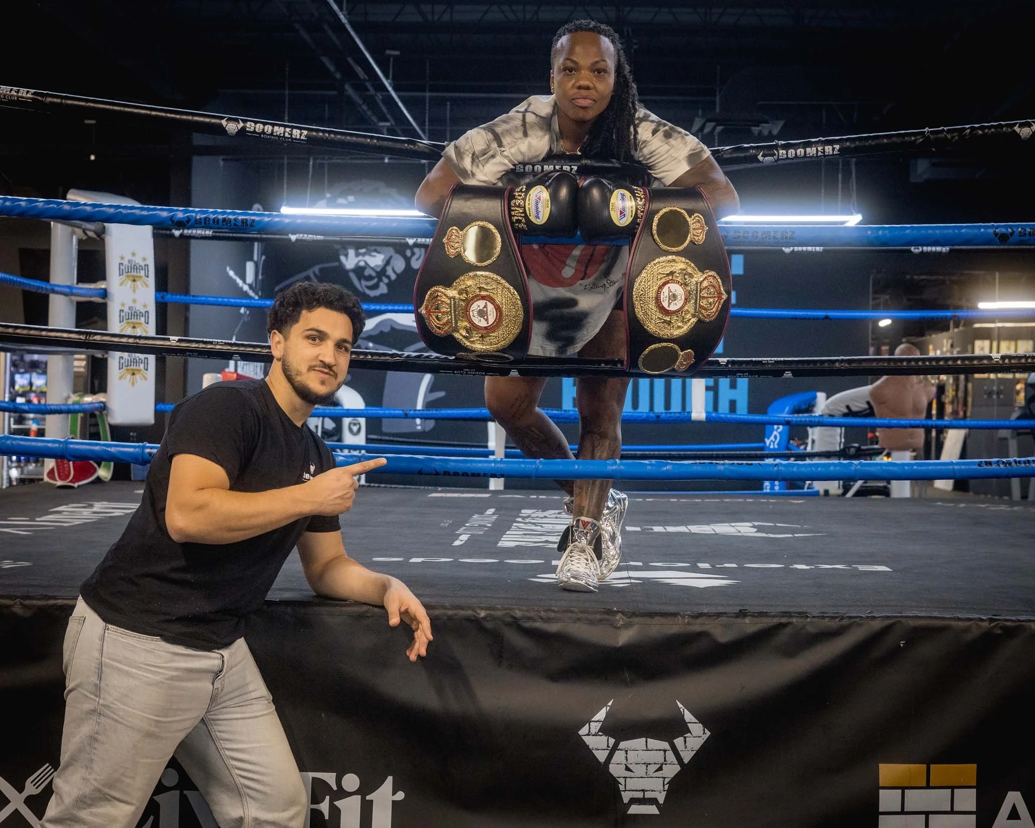 A female boxer with braided hair leaning on the top rope of a boxing ring, with championship belts hanging over her shoulders, and a serious expression. Two men are in front of the ring, one pointing at her and the other smiling. The background featu