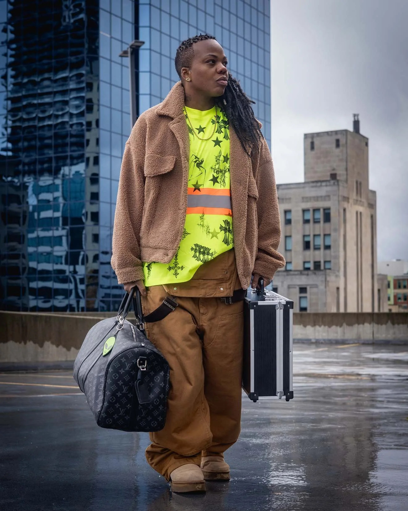 A person standing outdoors in an urban setting with tall glass buildings and concrete structures, wearing a yellow-green graphic T-shirt, a brown teddy coat, brown pants, and holding a black suitcase and a travel bag.