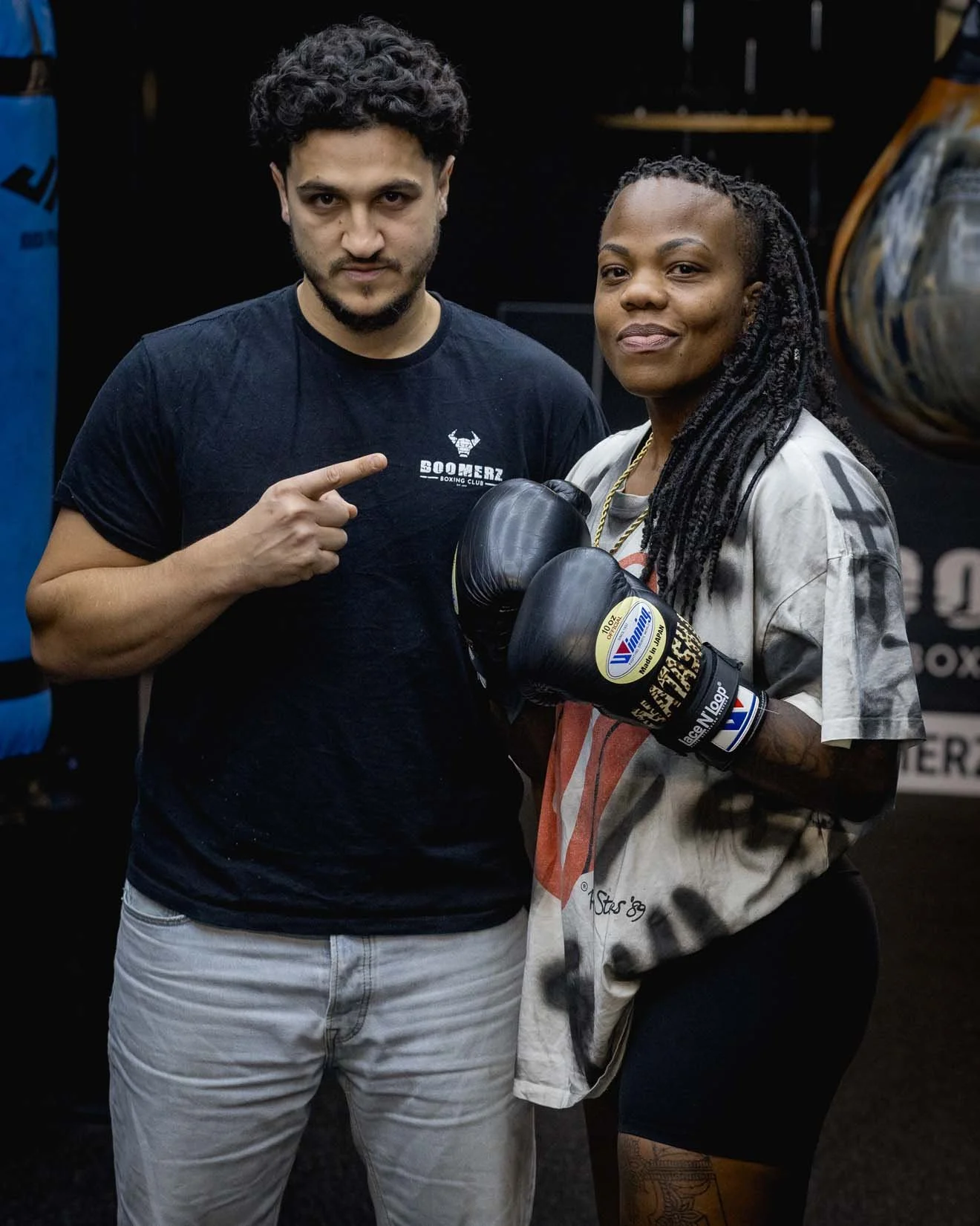 A man and woman posing together in a boxing gym. The man is pointing at the woman, who is holding boxing gloves. The woman has long, dreadlocked hair and is wearing a t-shirt and boxing gloves. The man is wearing a black t-shirt and light-colored pan