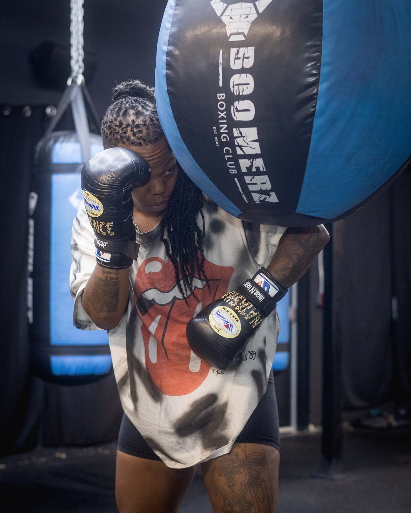A woman practicing boxing in a gym, wearing boxing gloves and punching a large blue and black punching bag.
