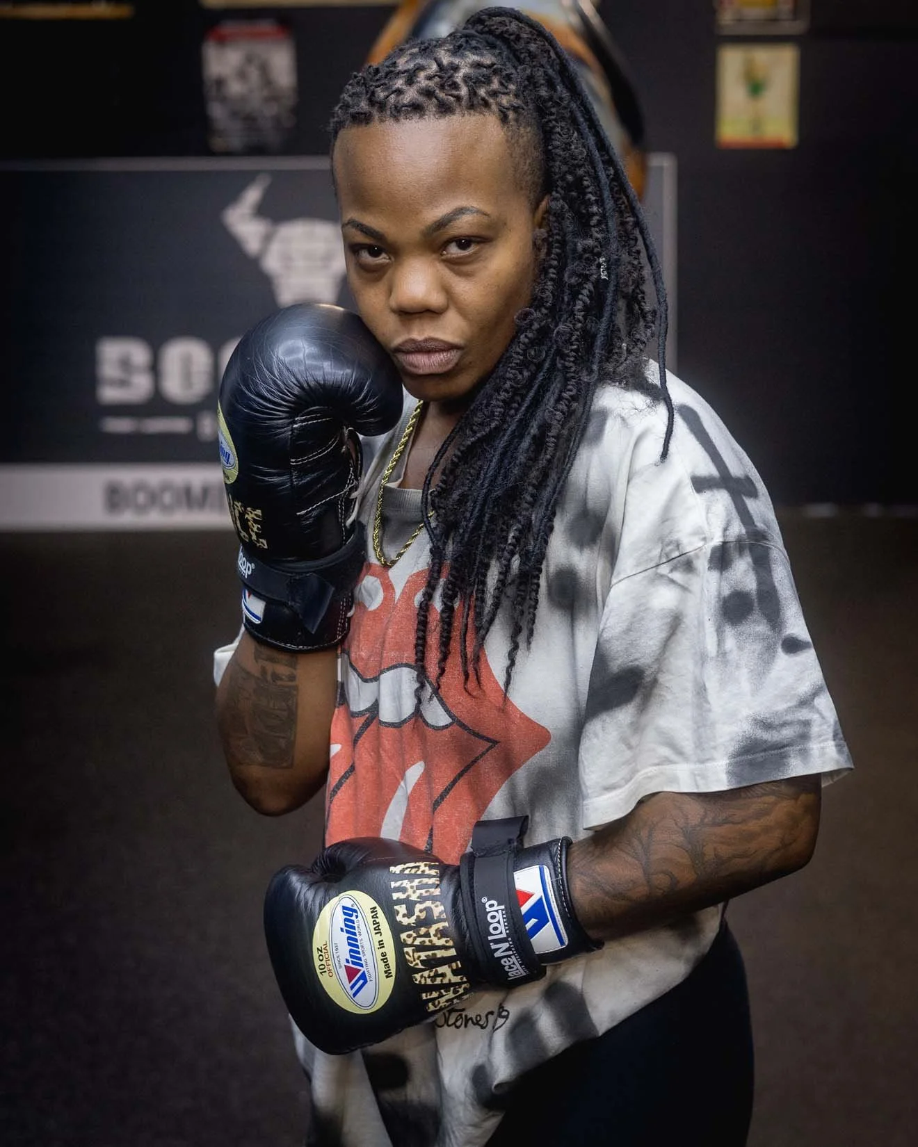 A woman with dreadlocks wearing boxing gloves, a graphic t-shirt, and tattoos on her arms, posing in a boxing gym.