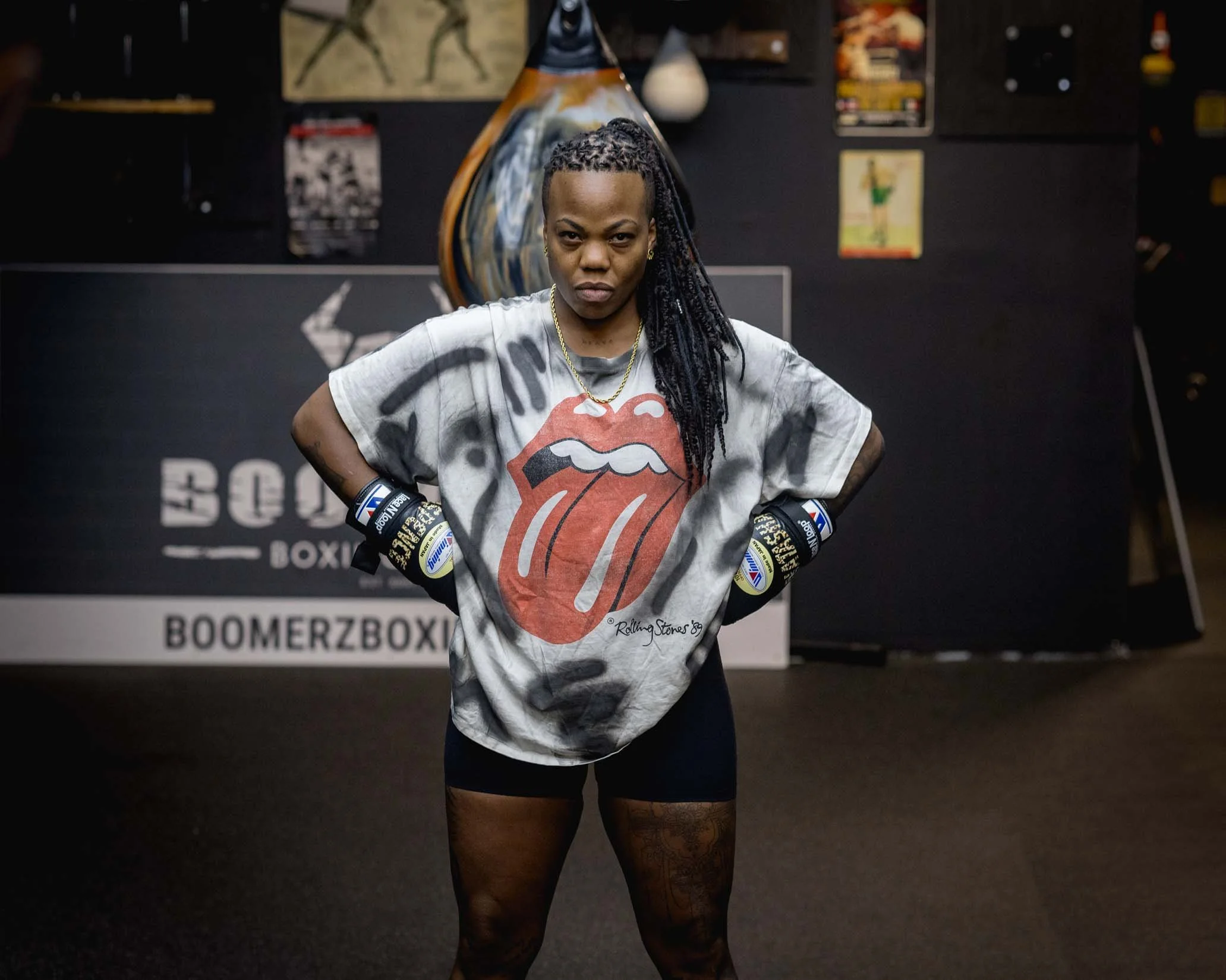 A woman standing in a gym with boxing gloves, wearing a Rolling Stones t-shirt, with dark hair in braids, in front of a wall with posters and fitness equipment.