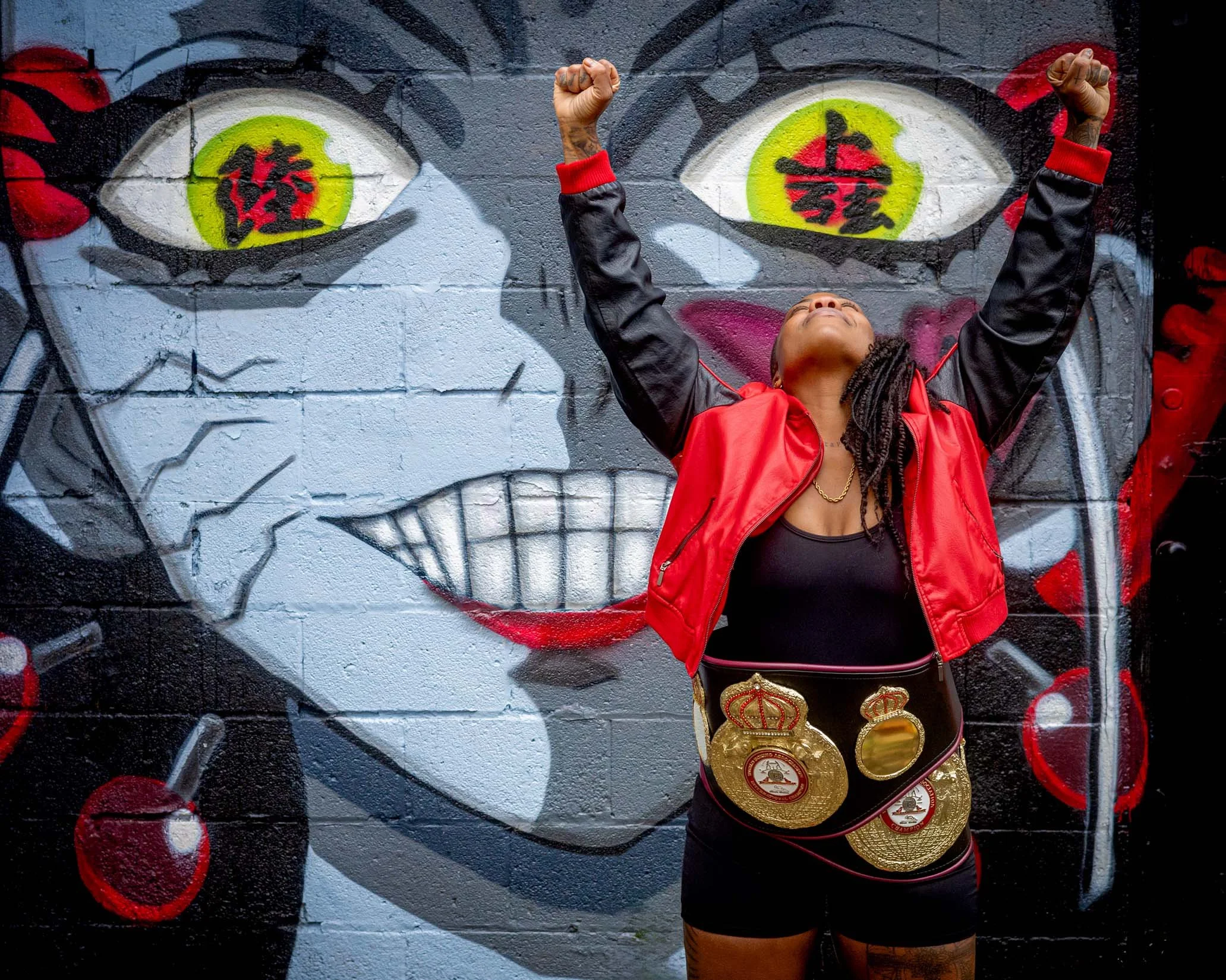 A woman standing in front of a graffiti wall, wearing a championship boxing belt around her waist and raising her fists in the air.