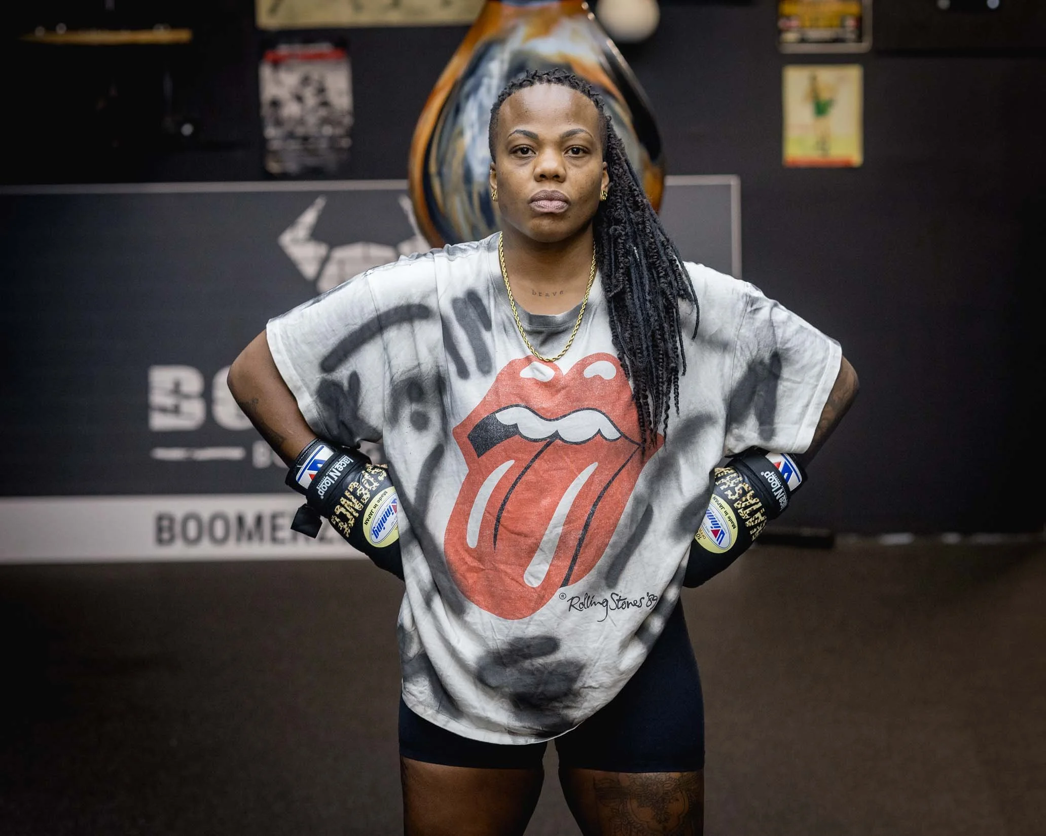 A woman with braided hair and wearing a Rolling Stones T-shirt stands with her hands on her hips in a gym, with boxing gloves on her hands, in front of a dark background with posters and equipment.