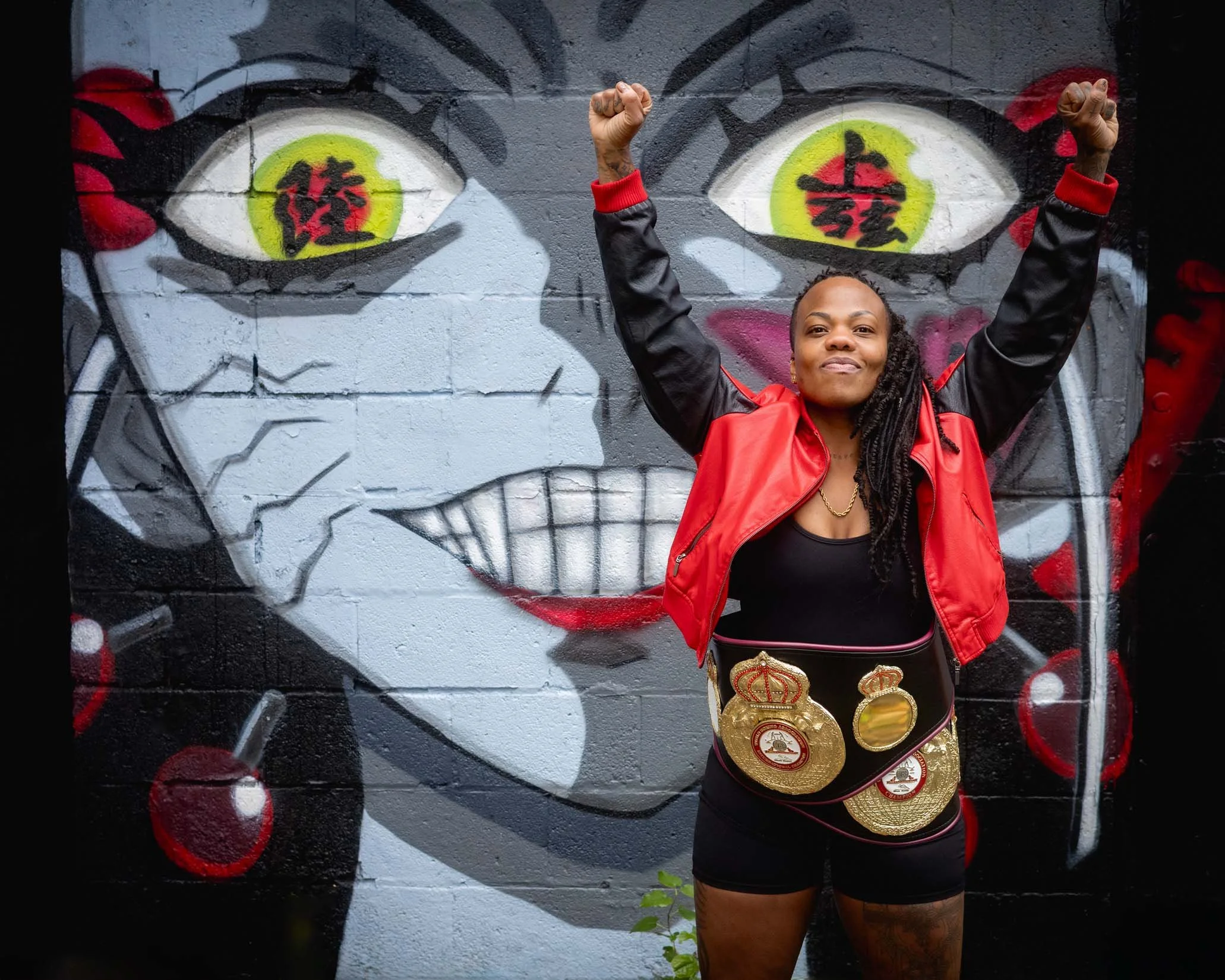 A woman standing in front of graffiti art on a wall with her arms raised in a triumphant pose. She is wearing a black shirt, a red jacket, and multiple gold championship belts around her waist. The graffiti art behind her depicts a large eye with bol