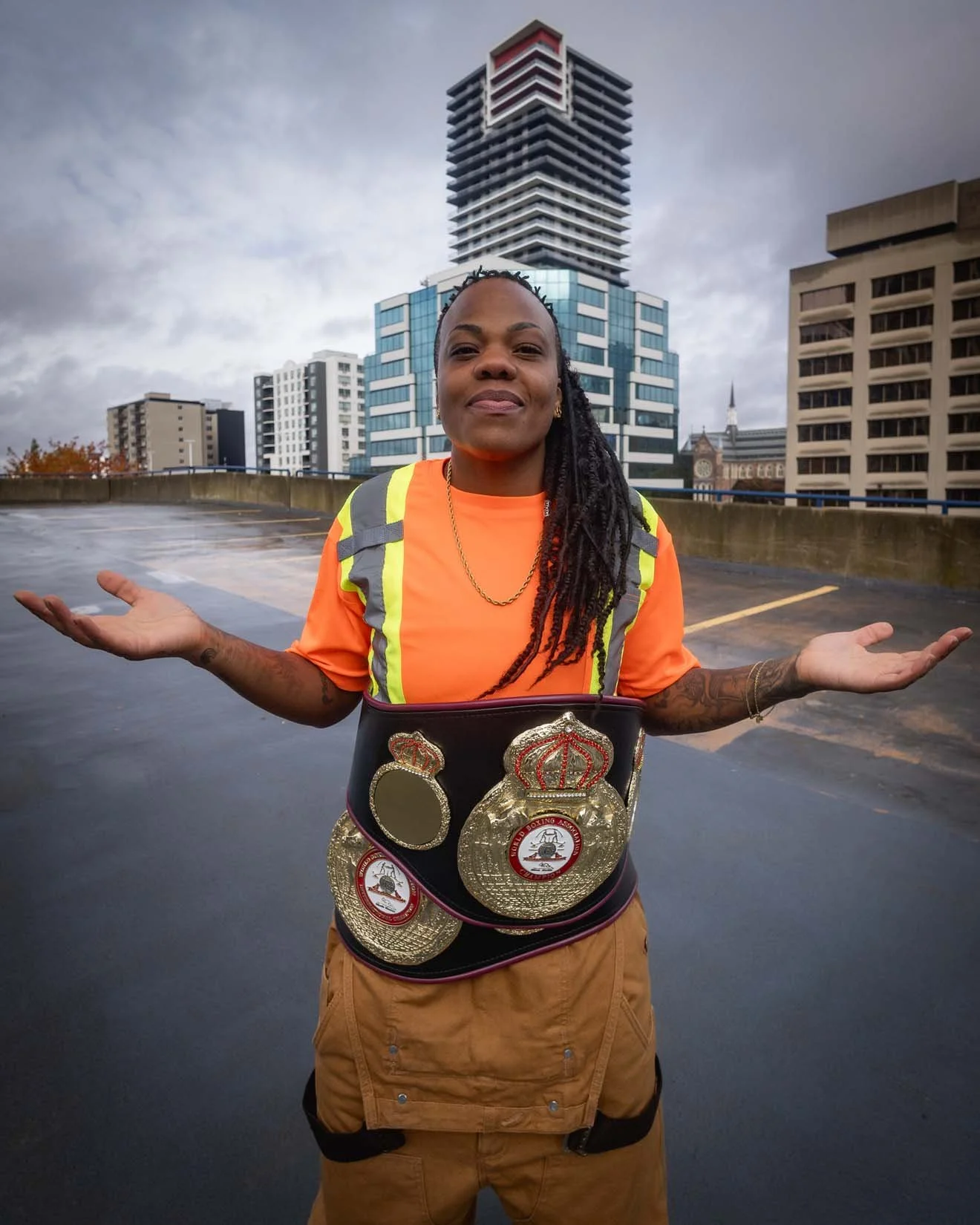 A woman wearing a boxing championship belt and an orange safety vest stands on a rooftop parking lot with city buildings in the background, shrugging her shoulders.