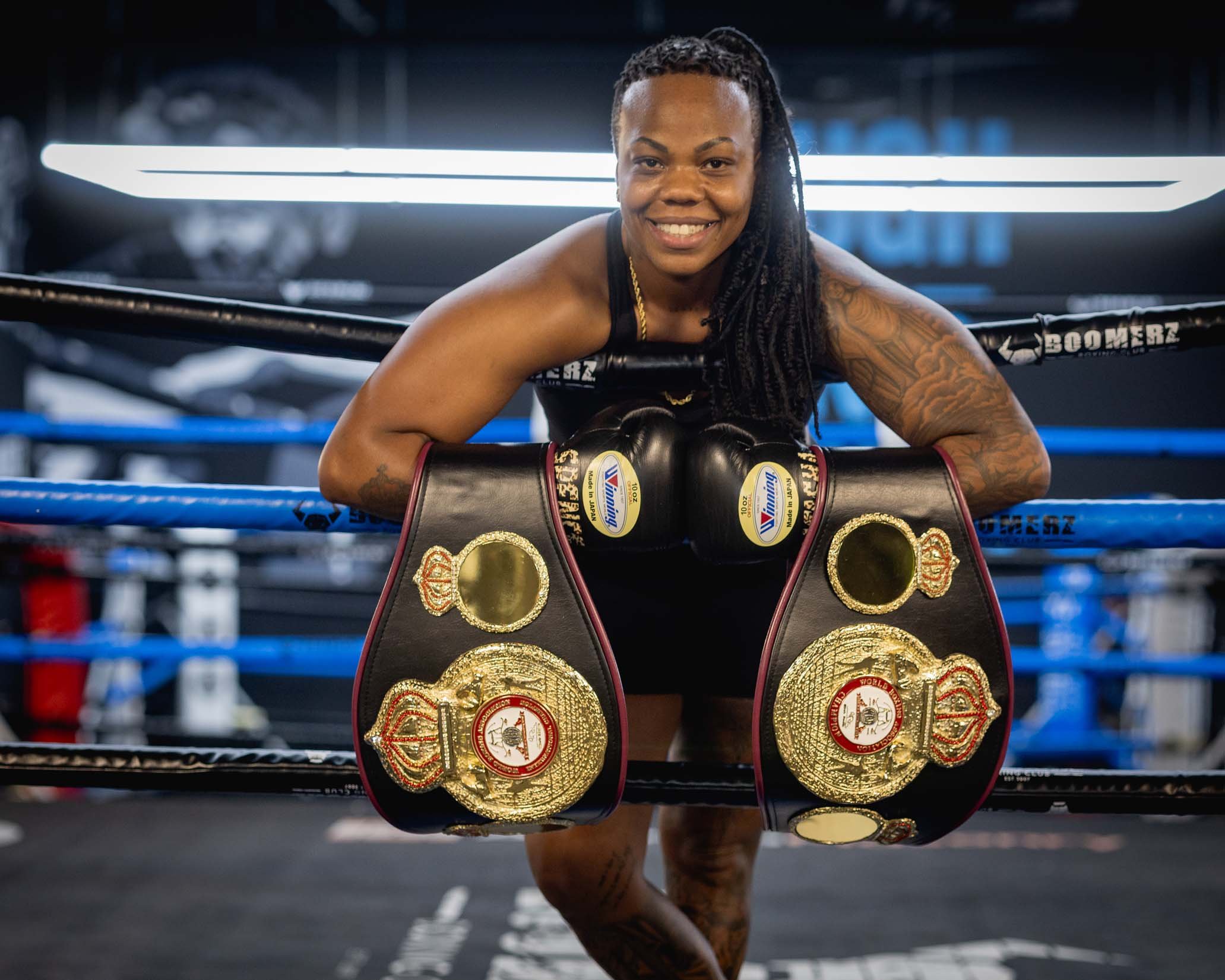 A female boxer leaning on the ropes of a boxing ring, smiling, with multiple championship belts hanging in front of her.