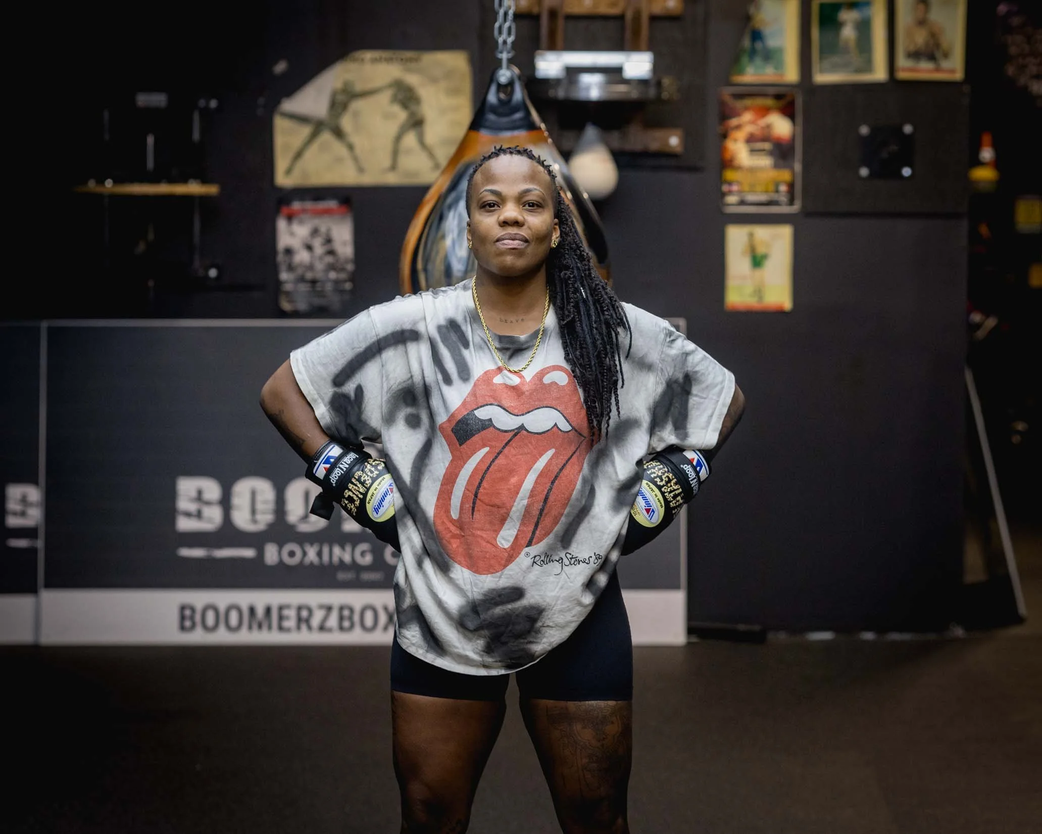 A woman with long braids stands in a boxing gym, wearing a Rolling Stones T-shirt, black shorts, and boxing gloves, with tattoos on her legs and a confident expression. The background features boxing posters and a punching bag.