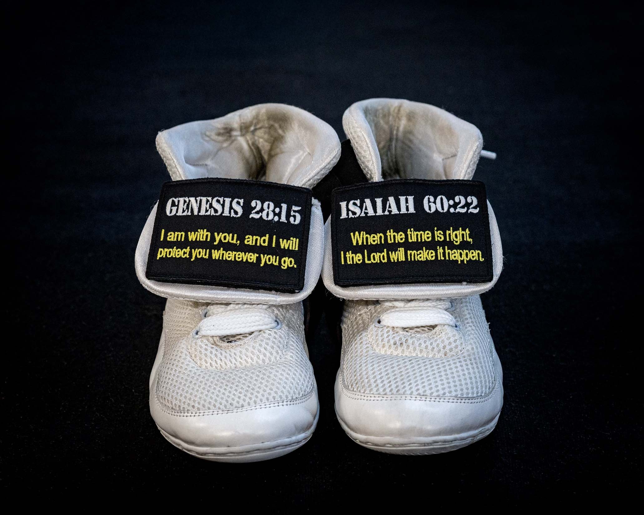 A pair of white athletic shoes with black labels on the tongues, printed with Bible verses Genesis 28:15 and Isaiah 60:22, resting on a black surface.