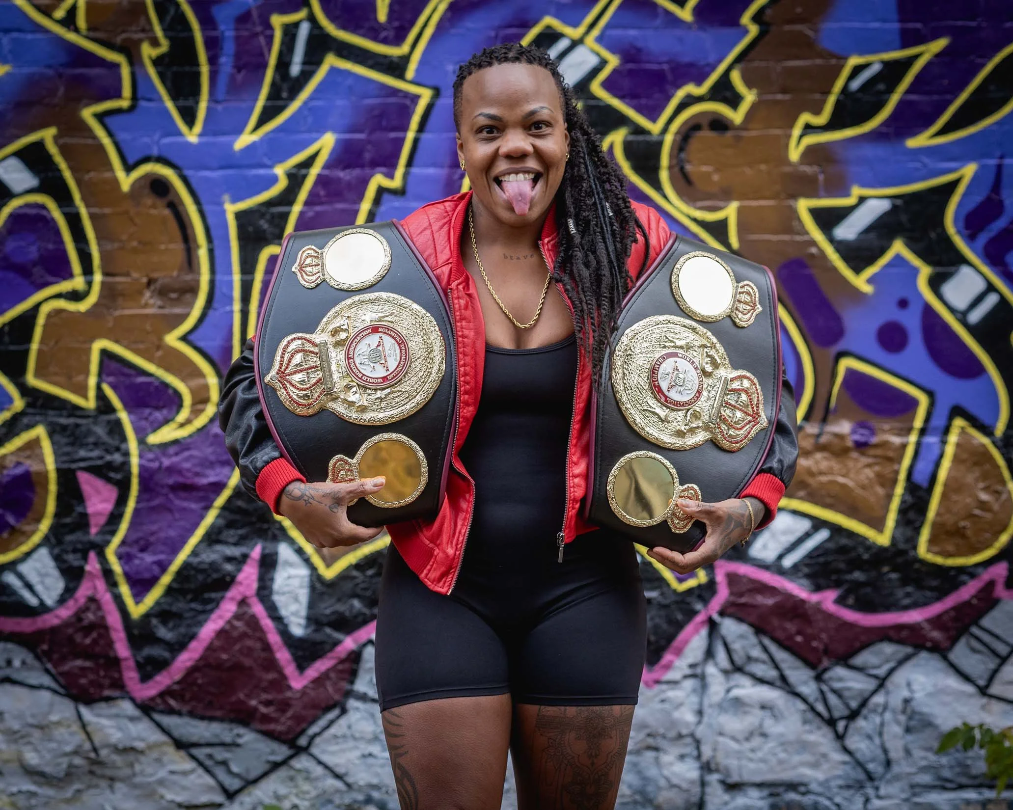 A woman in a red jacket and black outfit with tattoos on her legs sticking her tongue out and holding two championship belts, standing in front of a colorful graffiti wall.