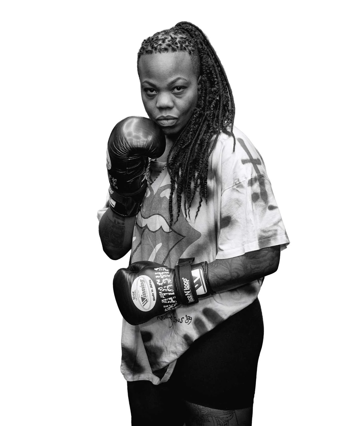 A woman with dreadlocks wearing a sports t-shirt and boxing gloves, posed in a fighting stance against a plain background.