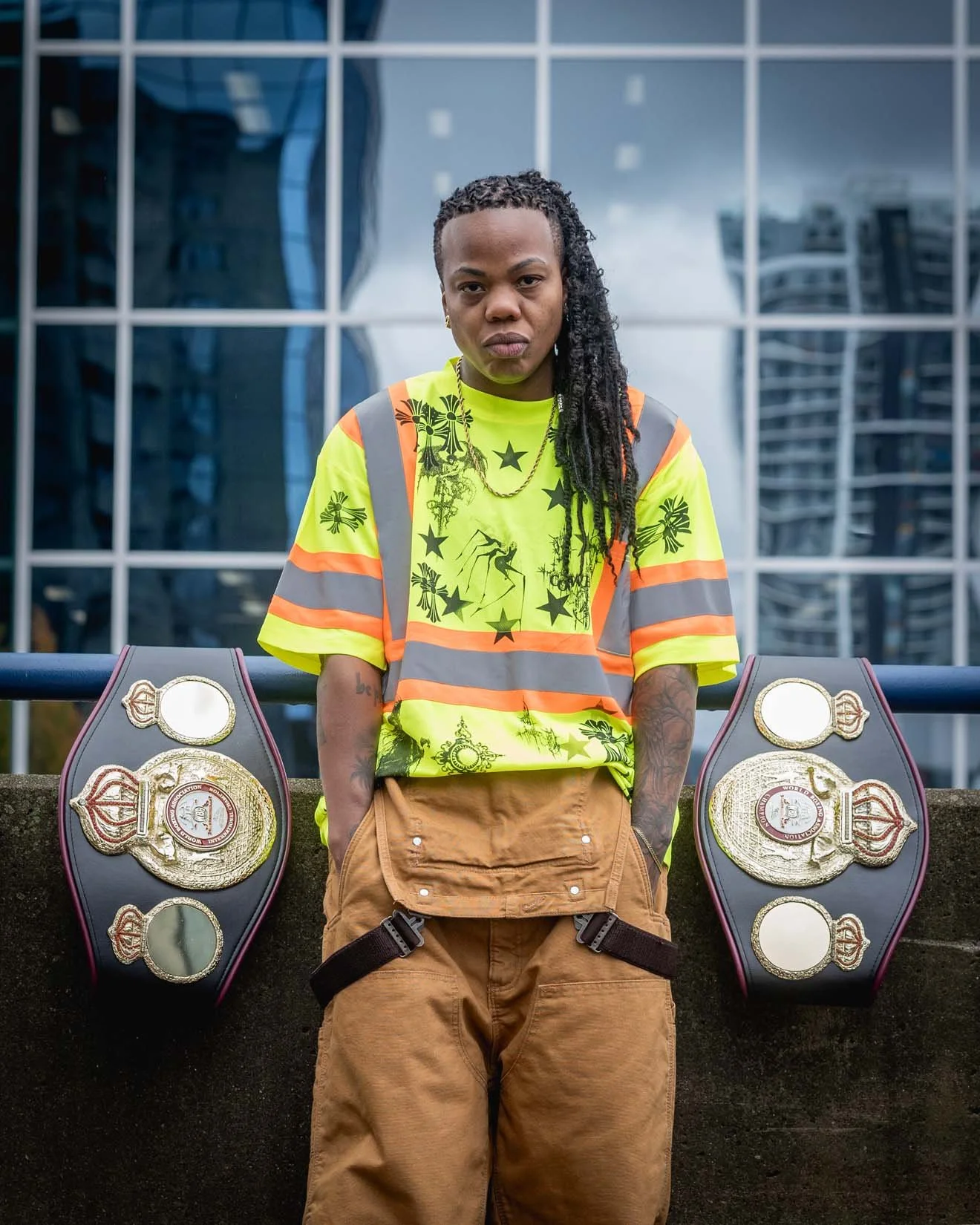 A person with long dreadlocks standing outdoors in front of a glass building, wearing a neon yellow safety vest over a graphic t-shirt, tan cargo pants, and displaying two championship belts on either side.