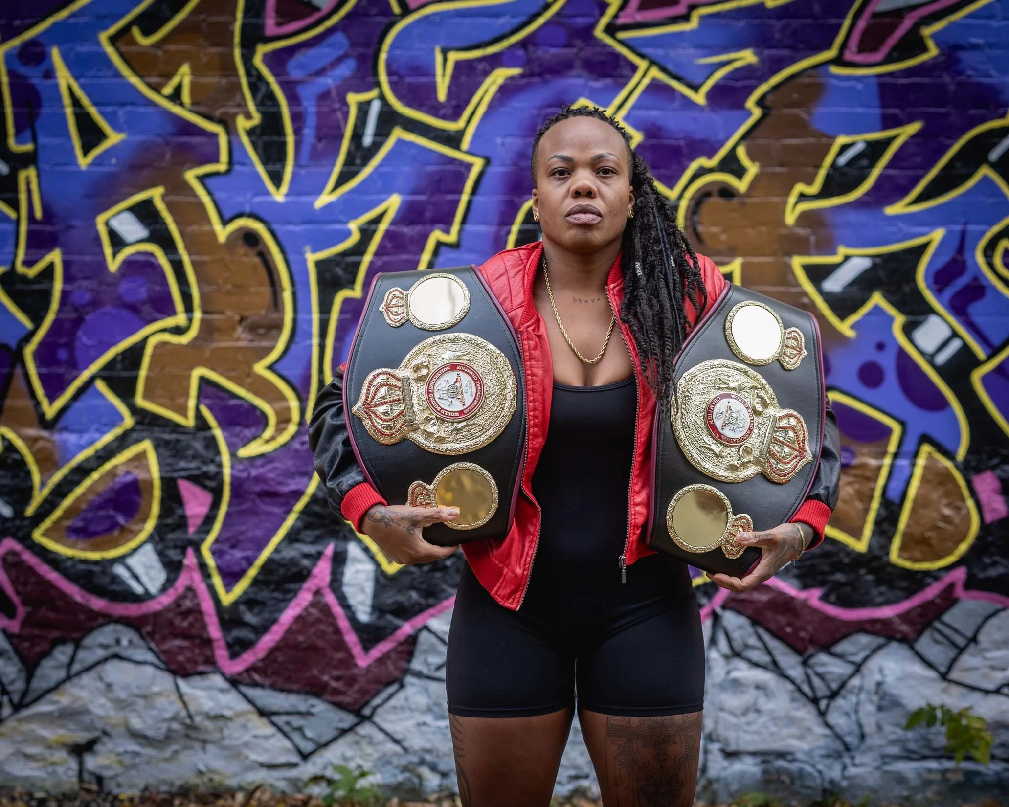 A woman wearing a red jacket and black outfit, holding multiple championship belts, standing in front of a colorful graffiti mural on a brick wall.