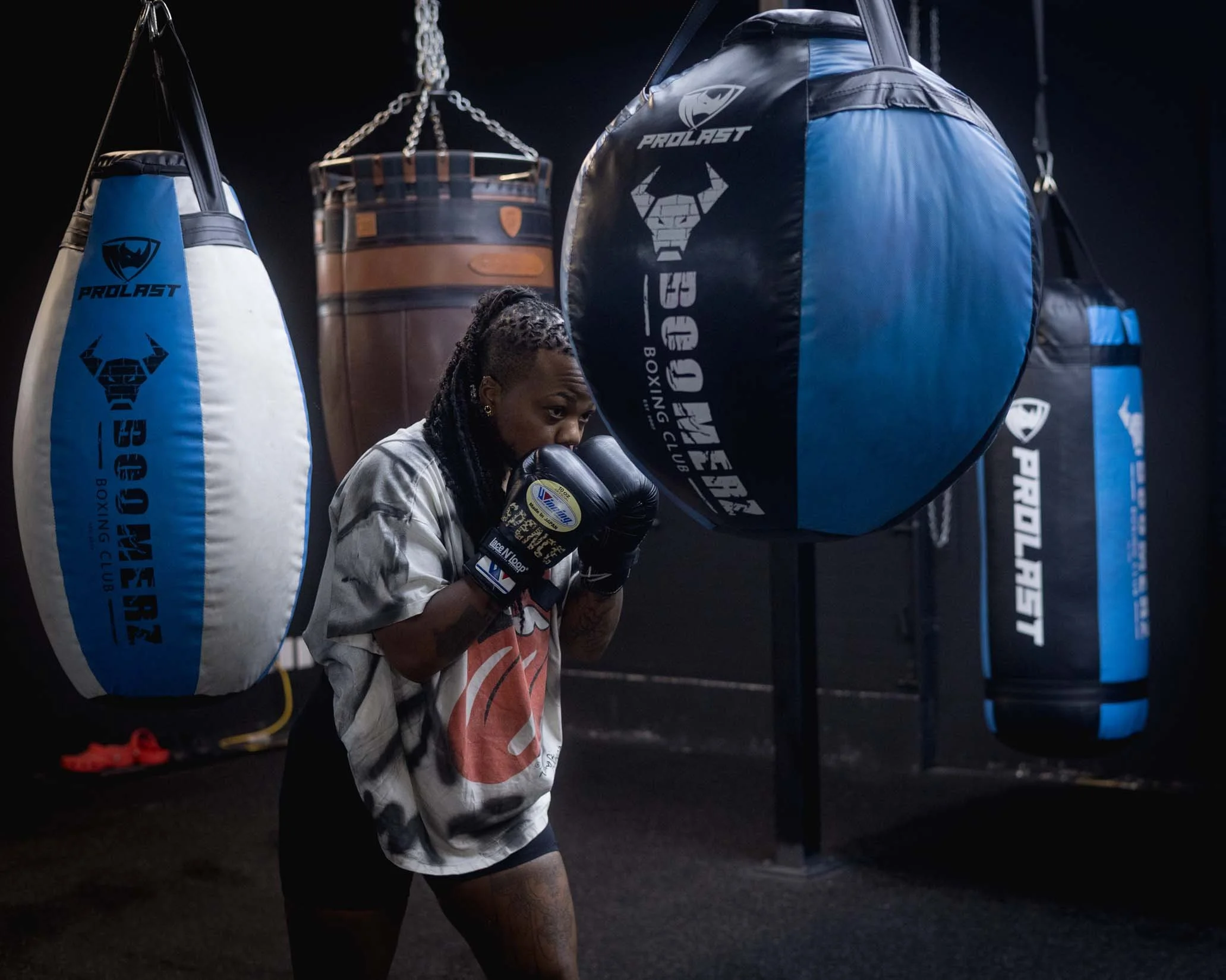 A woman wrapping her hands with black boxing hand wraps in a gym, standing near blue and black punching bags.