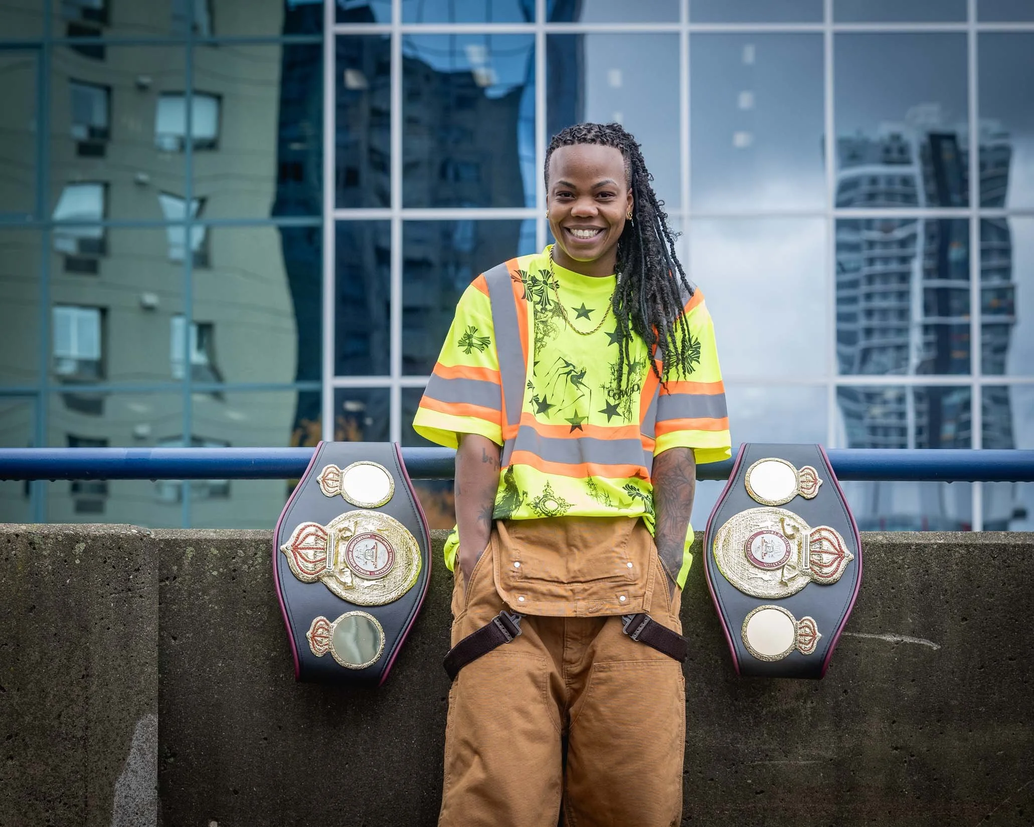 A smiling woman in a yellow safety vest and brown pants standing behind a concrete barrier with championship belts on either side, with a modern glass building in the background.