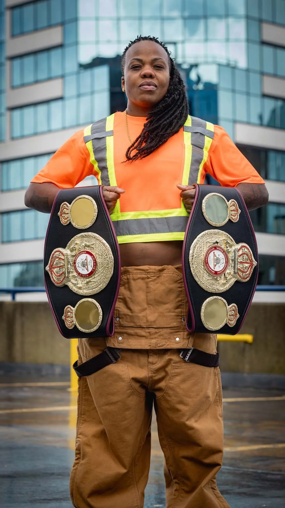 A woman in work attire holding two championship wrestling belts outside in front of a modern office building.