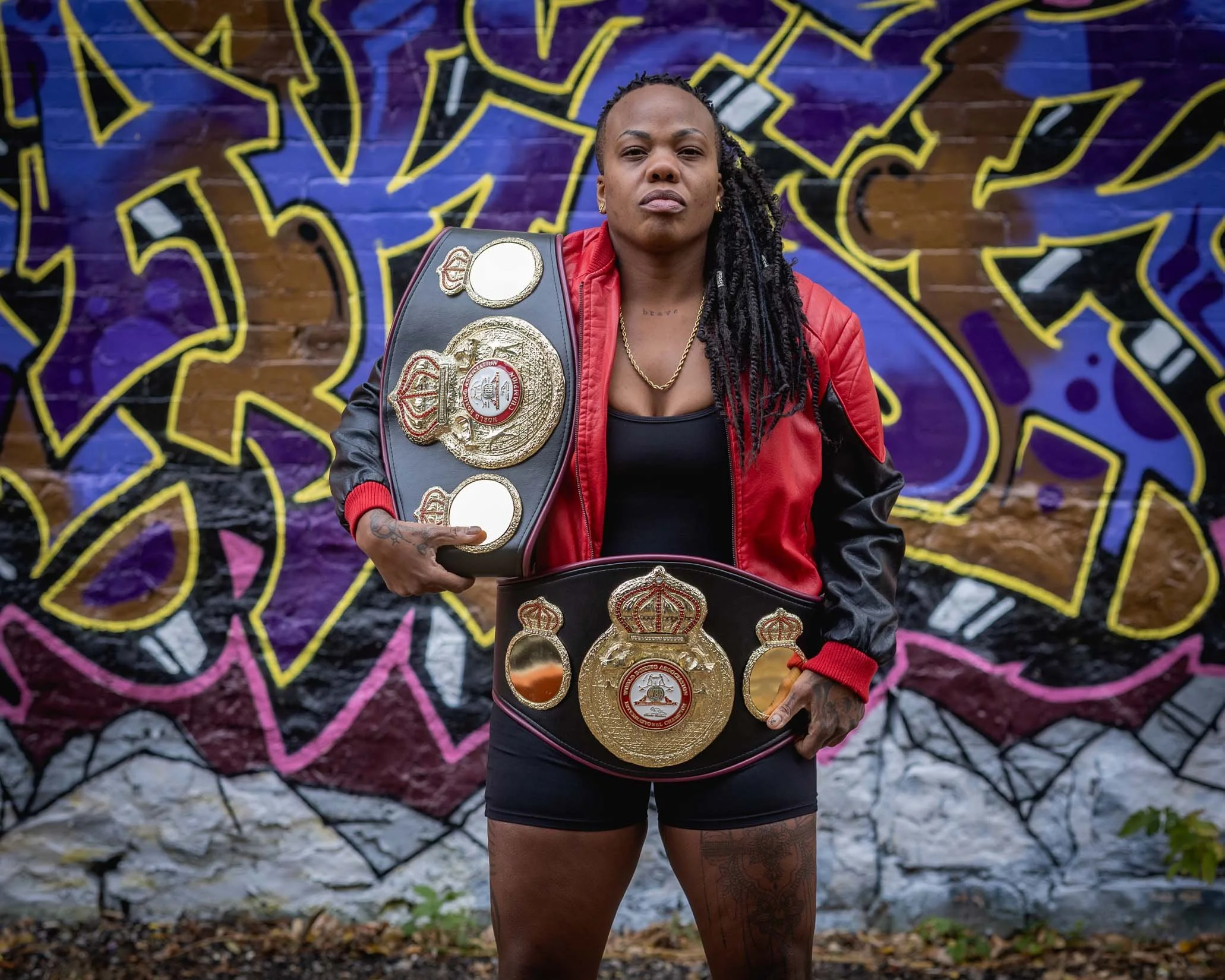Woman with long dreadlocks and tattoos wearing a red and black jacket, holding championship belts, standing in front of a graffiti wall.