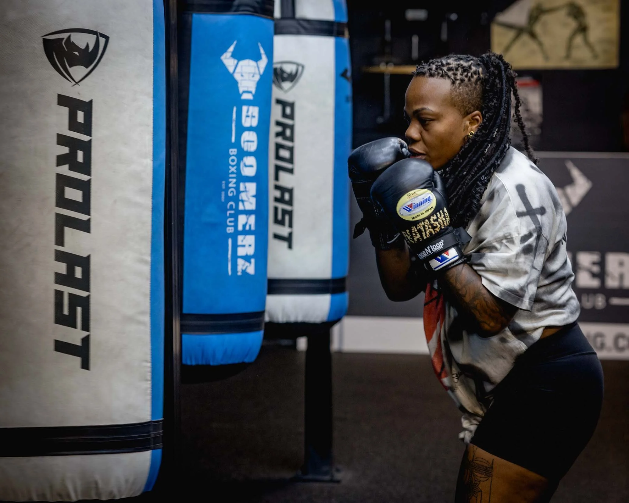 A woman with braided hair and tattoos on her arms wearing boxing gloves, practicing boxing at a training gym with punch bags.