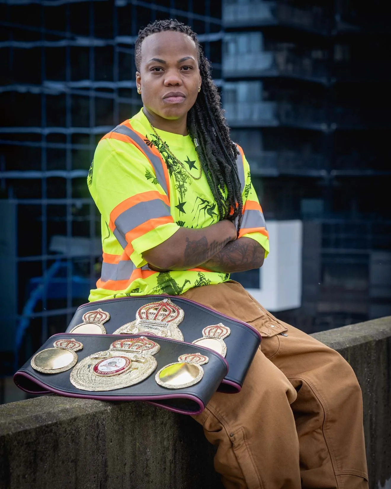 A woman with dreadlocks and tattoos, wearing a high-visibility safety vest, is sitting on a ledge with her arms crossed. In front of her is a display with championship belts and medals, and the background features a modern glass building.