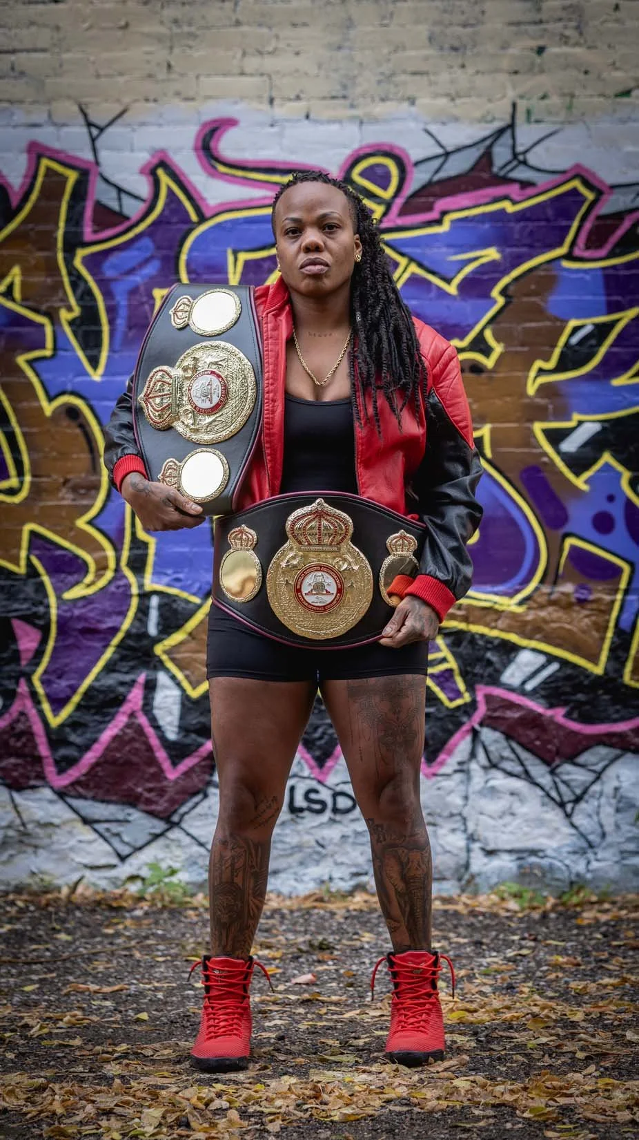 A woman holding two championship belts standing in front of a colorful graffiti wall.