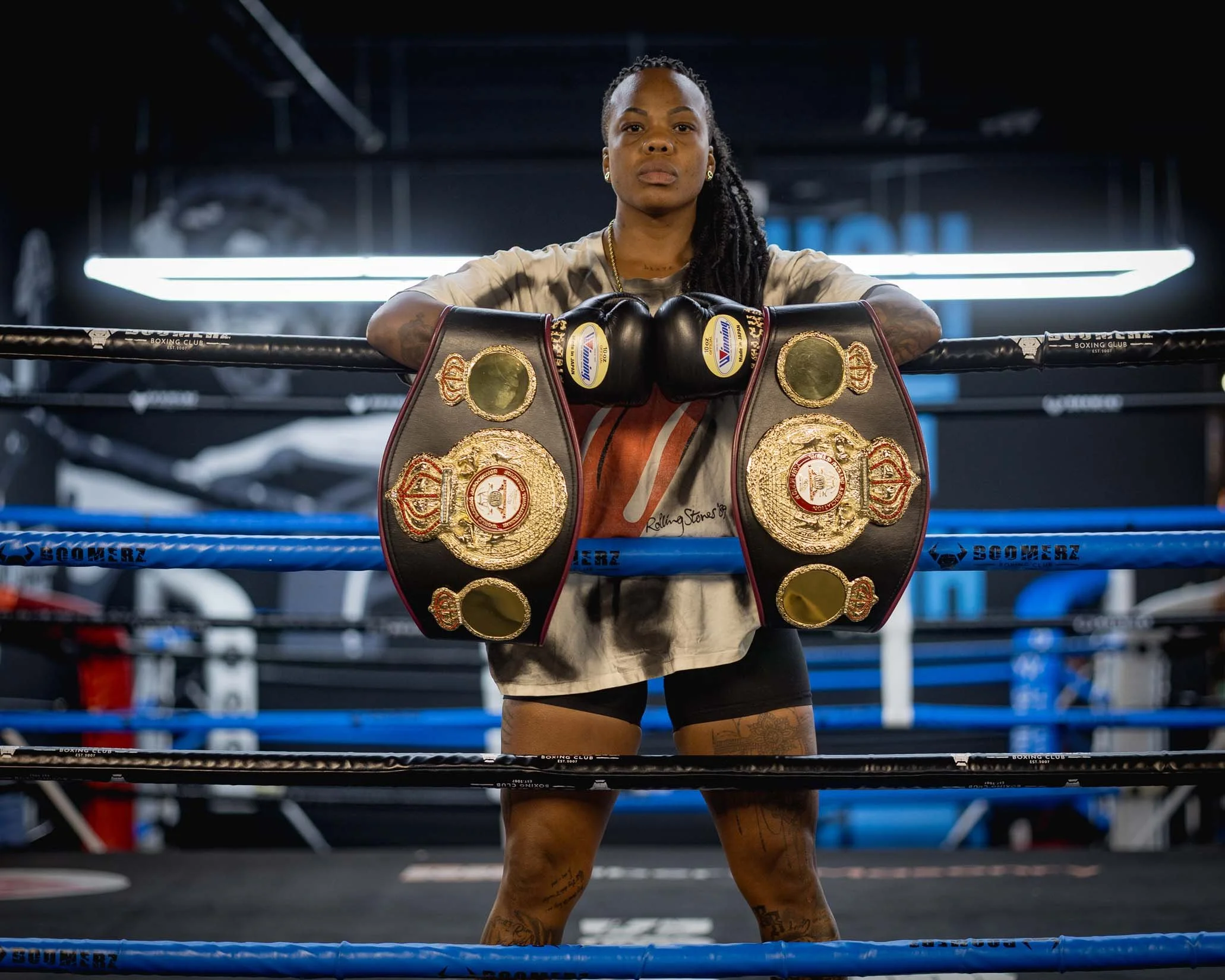 A female boxer in boxing gloves and championship belts standing in a boxing ring with her arms resting on the ropes, facing forward in a gym.