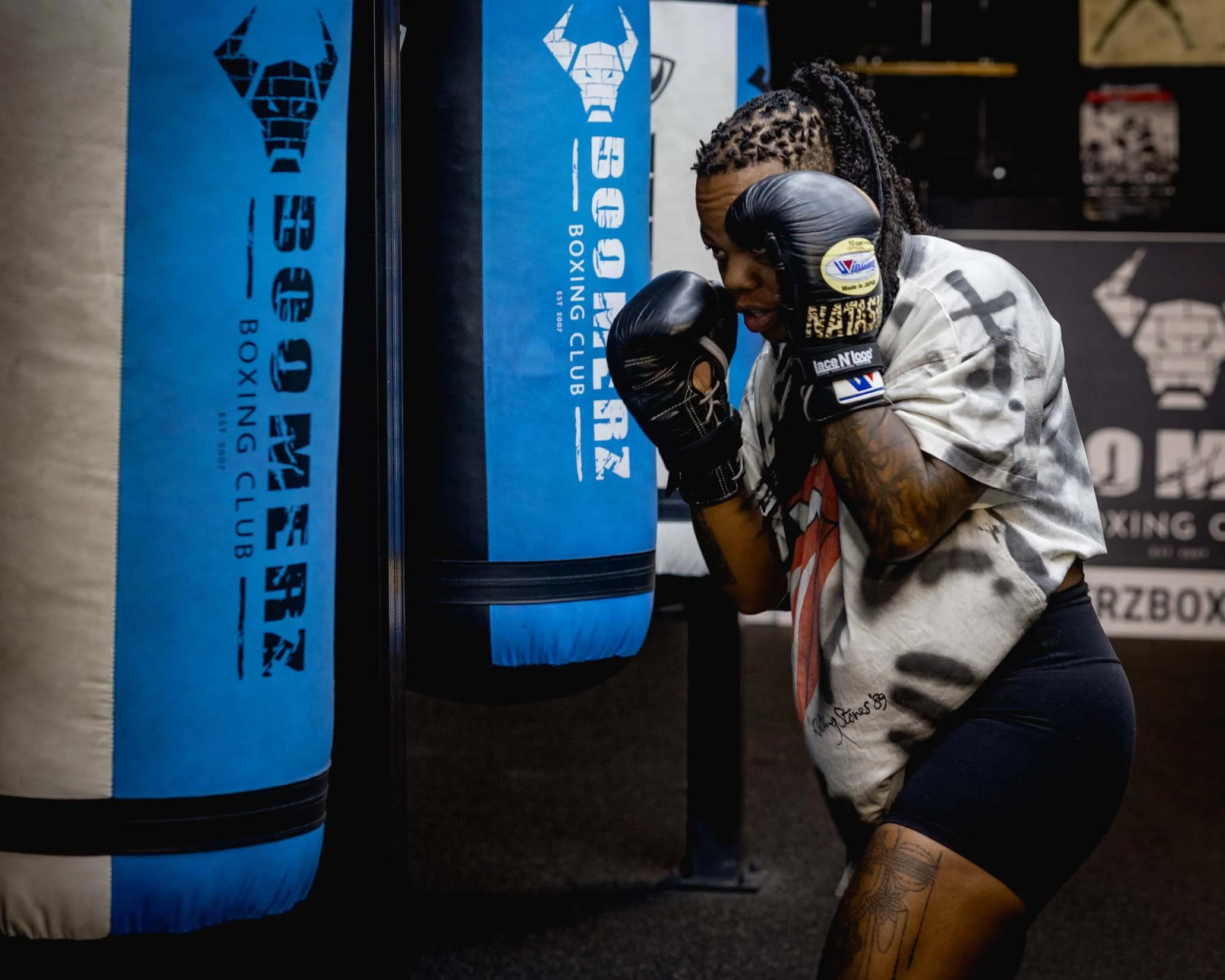 A woman with tattooed arms and dreadlocks, wearing boxing gloves and a boxing robe, is practicing boxing at a gym, hitting a blue punching bag with a tiger logo.