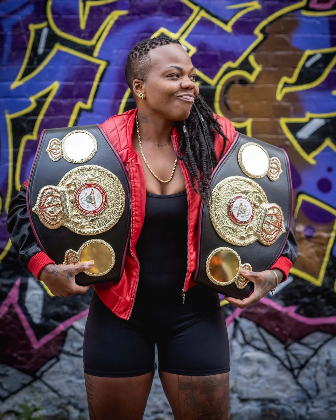 A woman with braided hair wearing a red jacket and black athletic wear, holding two championship belts with gold plates, stands in front of graffiti art on a brick wall.