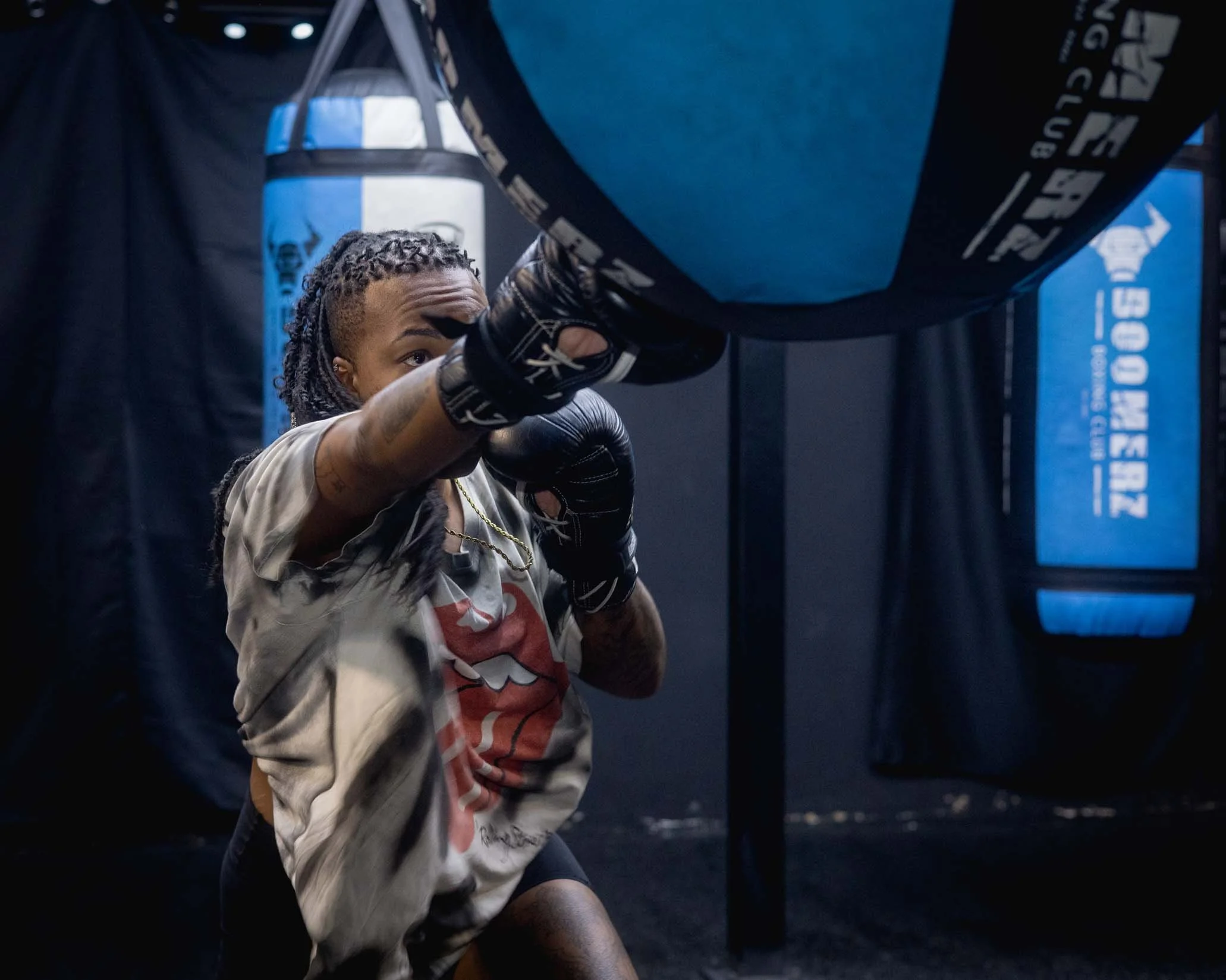 A person with dreadlocks wearing boxing gloves training with a punching bag in a gym.