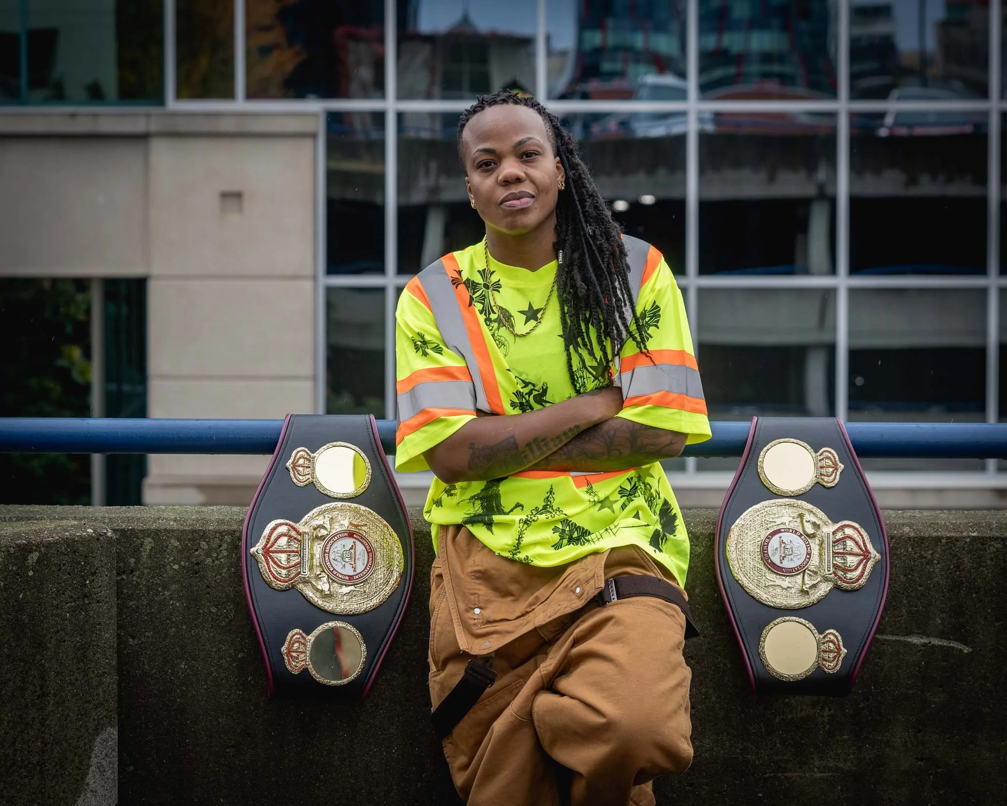 Woman with braids wearing a neon yellow safety shirt and tan shorts, standing outside with championship belts displayed in front of her.