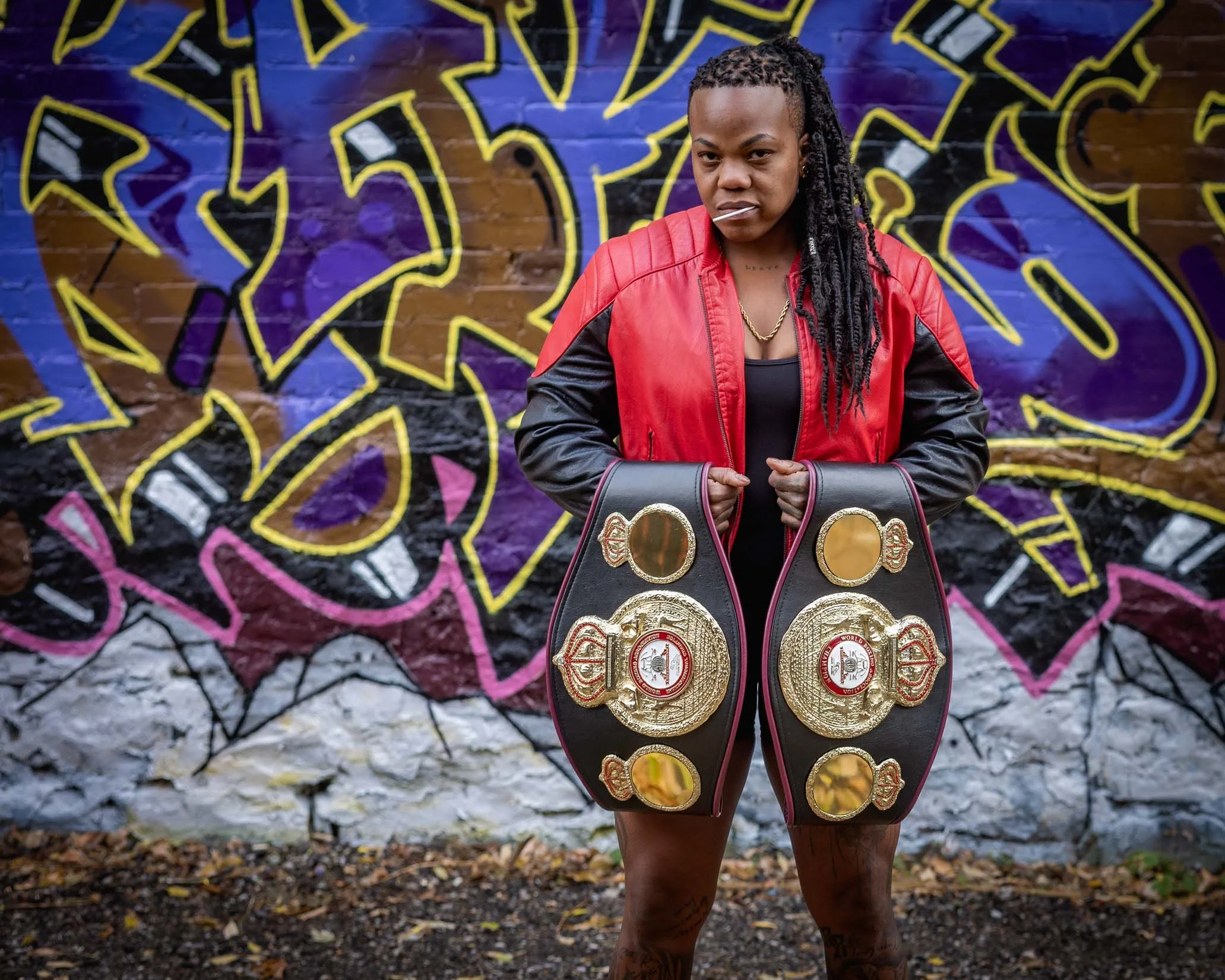 A woman with braided dreadlocks wearing a red and black jacket, holding multiple championship belts, standing in front of a colorful graffiti wall.